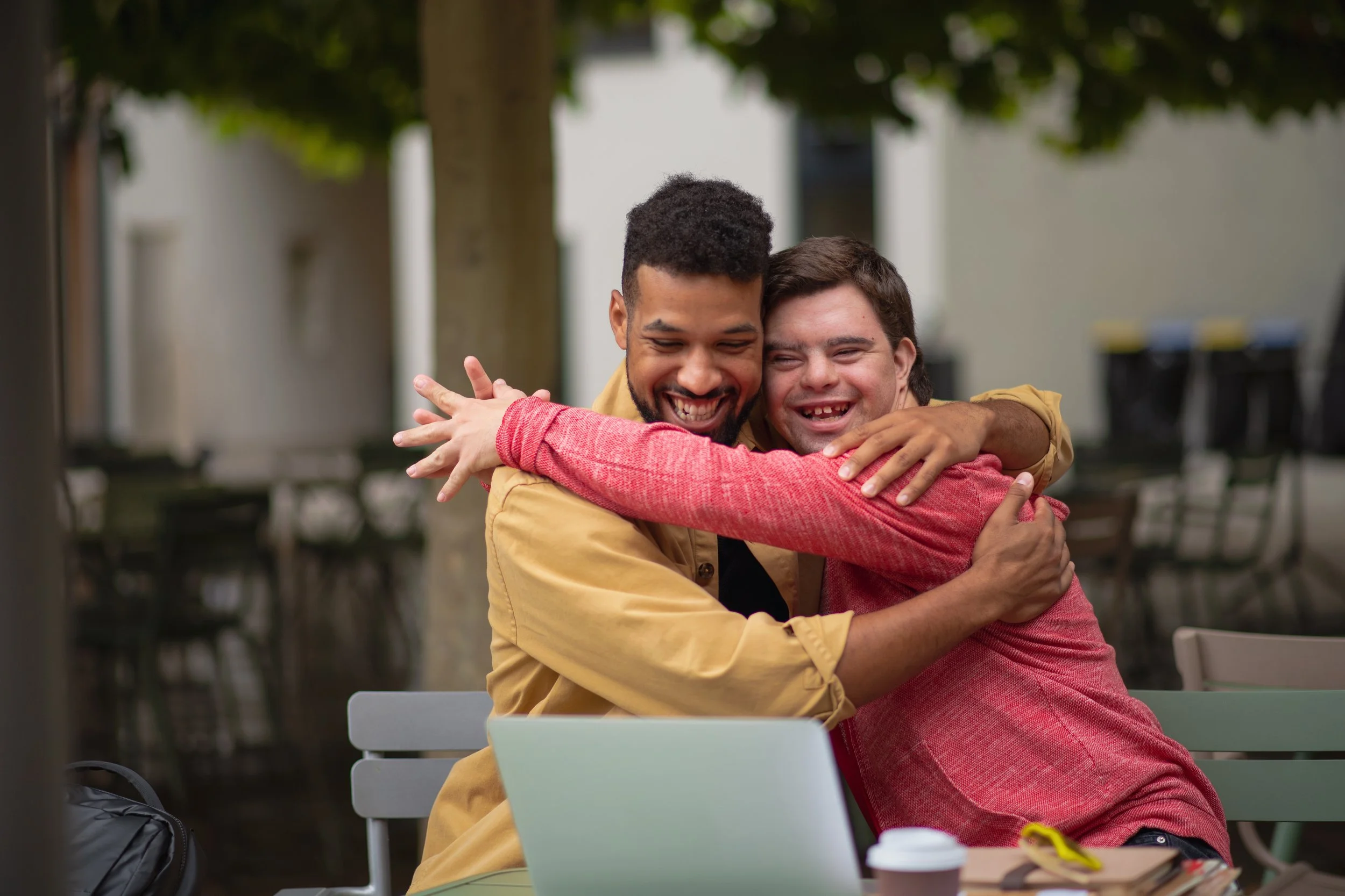 Two men sharing a warm hug and smiling at each other outdoors at a park or cafe, with trees and chairs in the background.
