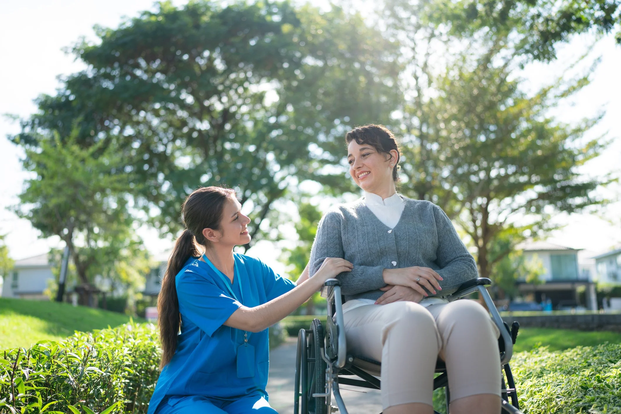 A nurse in blue scrubs kneeling next to an elderly woman in a wheelchair, outdoors with trees and houses in the background, both smiling at each other.