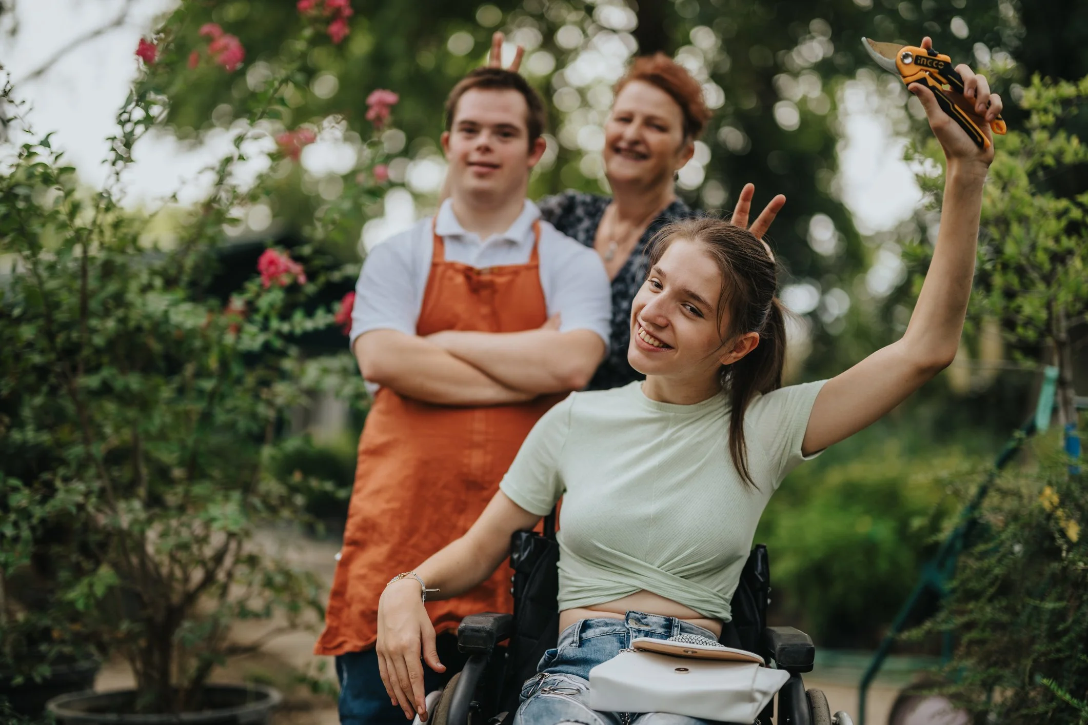 Three people, two women and one young man, smiling and making peace signs behind a woman in a wheelchair taking a selfie in a garden with green bushes and pink flowers.