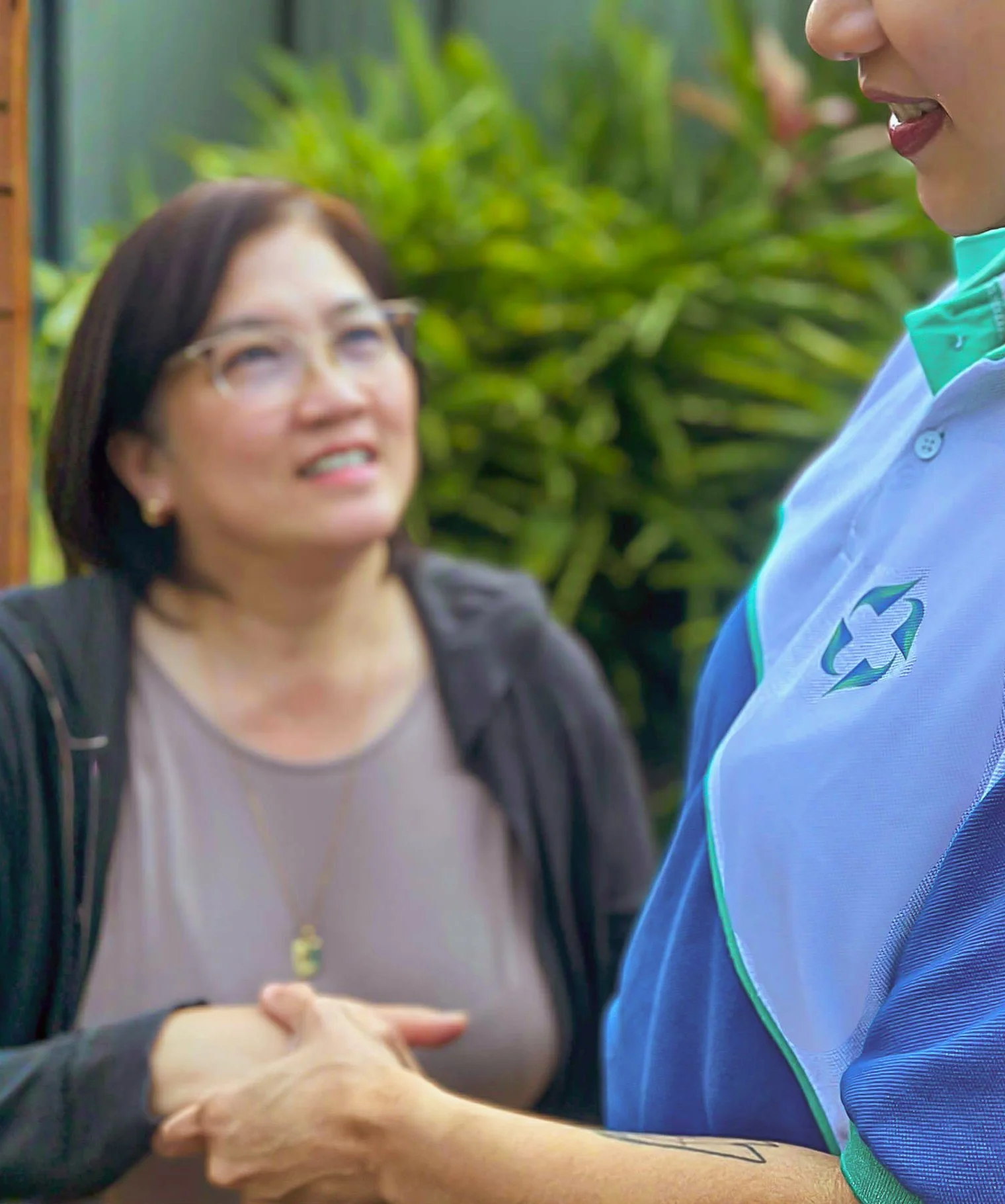 A woman and a person in a uniform shaking hands outdoors with lush green plants in the background.