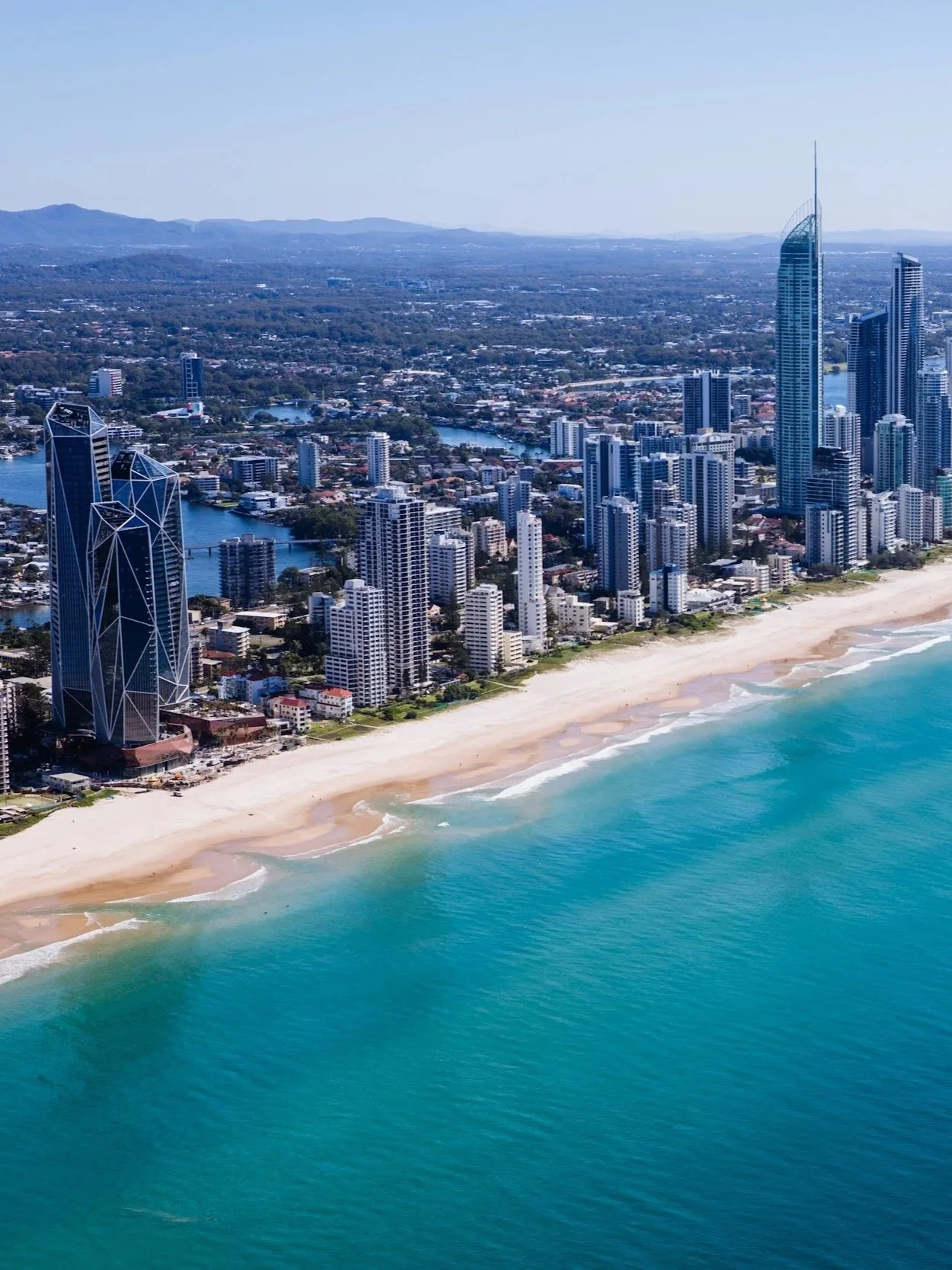 Aerial view of a coastal city with high-rise buildings along a sandy beach and turquoise ocean waves, with a river running through the city.