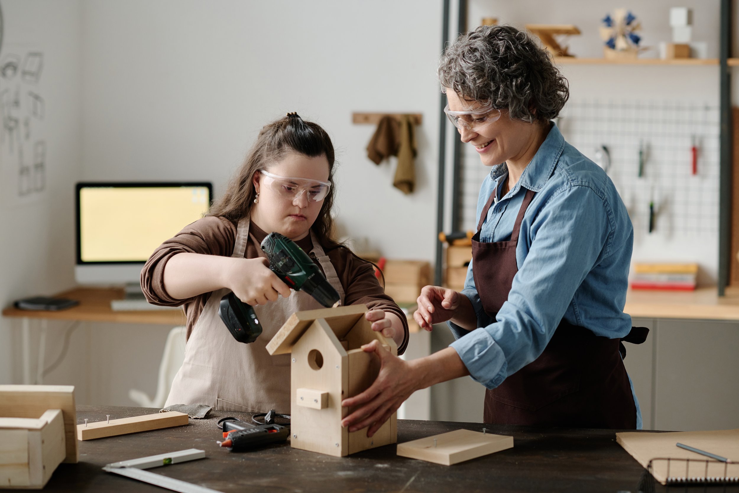 A woman and a girl working on building a wooden birdhouse in a woodworking workshop, both wearing safety glasses.