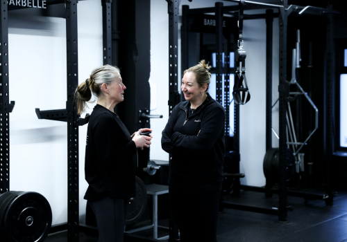 Two women talking and smiling in a gym with weightlifting equipment.