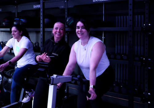 Three women smiling and biking at a fitness class in a gym with black storage shelves and barbells in the background.