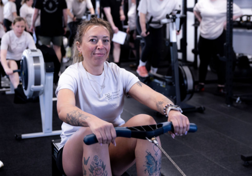 A woman sitting on a rowing machine at a gym, with gym equipment and people in the background.