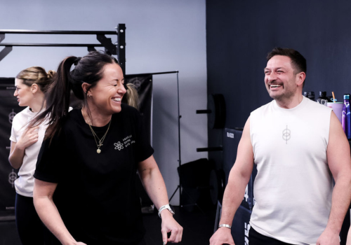 Two people smiling and laughing in a gym or fitness studio, with a woman in a black shirt and a man in a white sleeveless shirt.