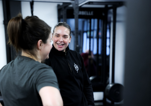 Two women smiling and talking at the gym, with fitness equipment in the background.