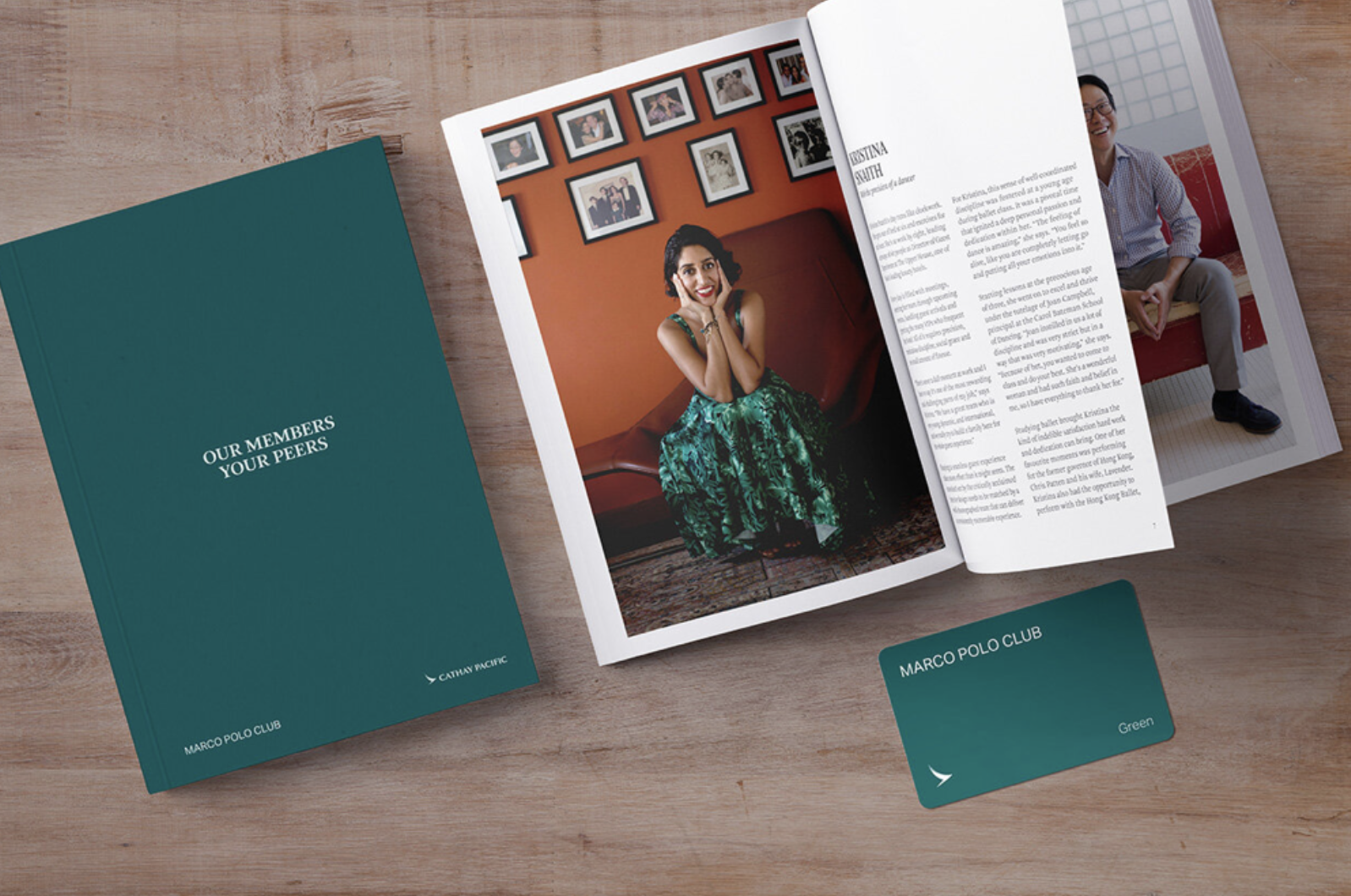 Open magazine showing a woman in a green dress sitting and smiling, with framed photos on the wall behind her, on a wooden surface alongside a teal booklet and a teal card from Marco Polo Club.