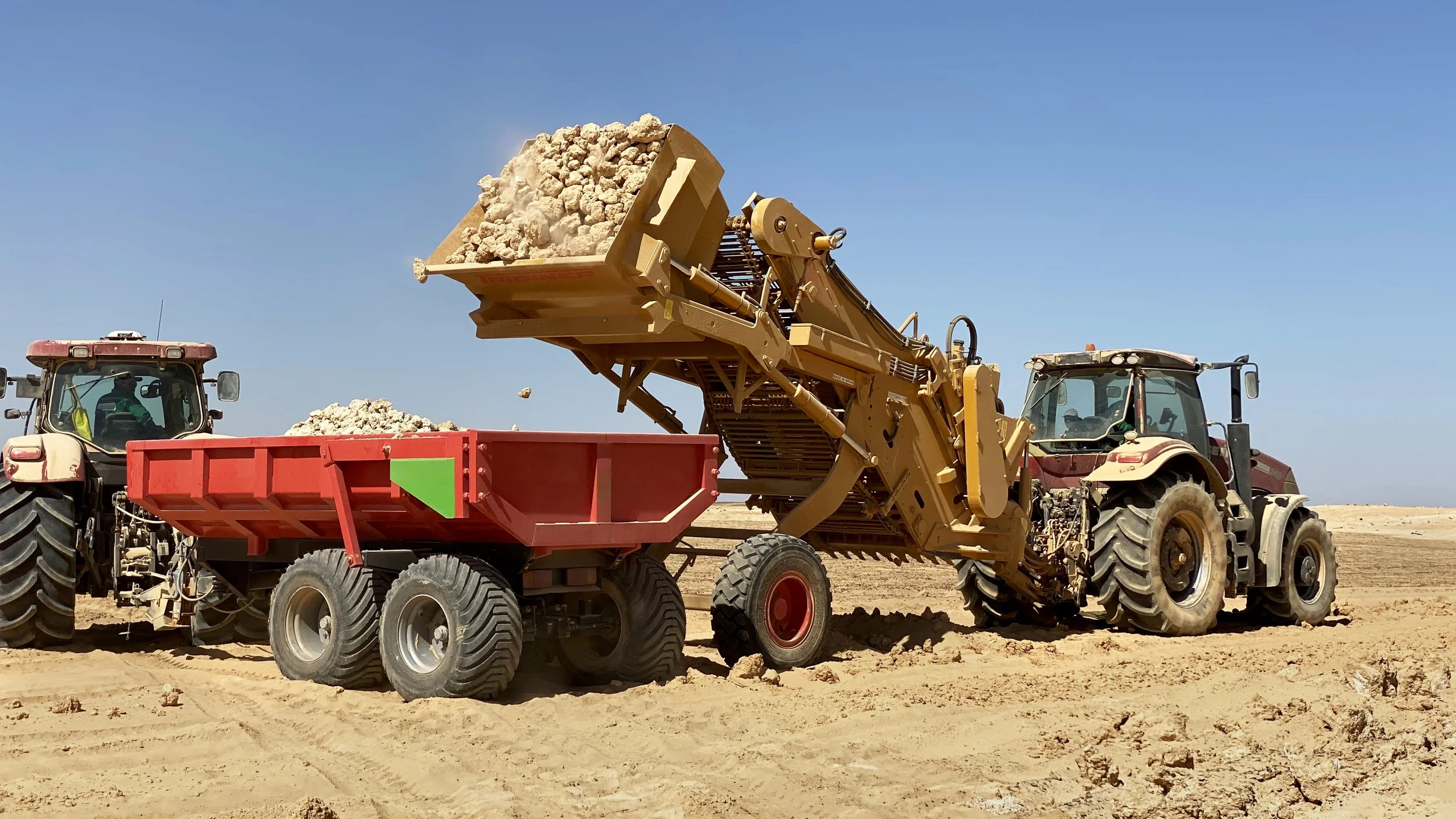 Dos tractores trabajando en un campo arenoso con maquinaria agrícola que carga tierra.