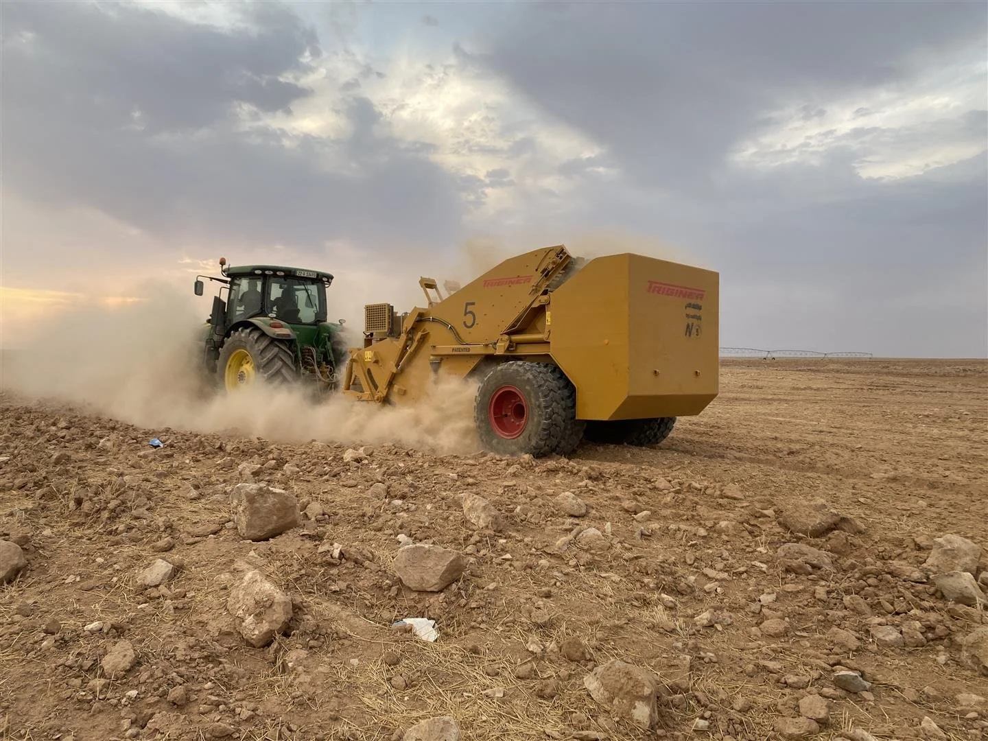 Una tractor amarilla está trabajando en un campo seco y arenoso bajo un cielo nublado durante el atardecer.