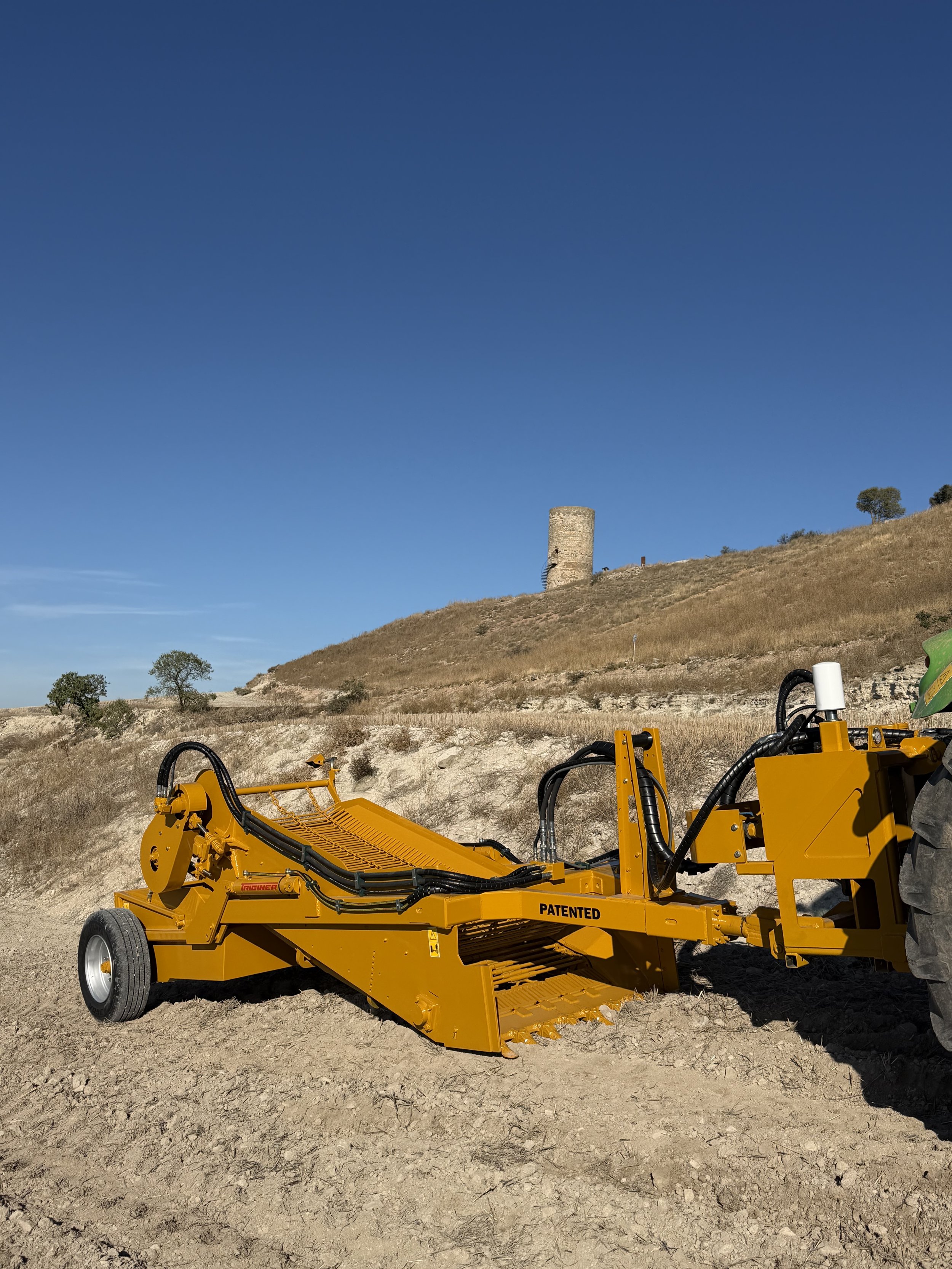 Maquinaria agrícola amarilla en un campo seco con colinas y una torre en el fondo. Máquina despedregadora Triginer trabajando el campo.