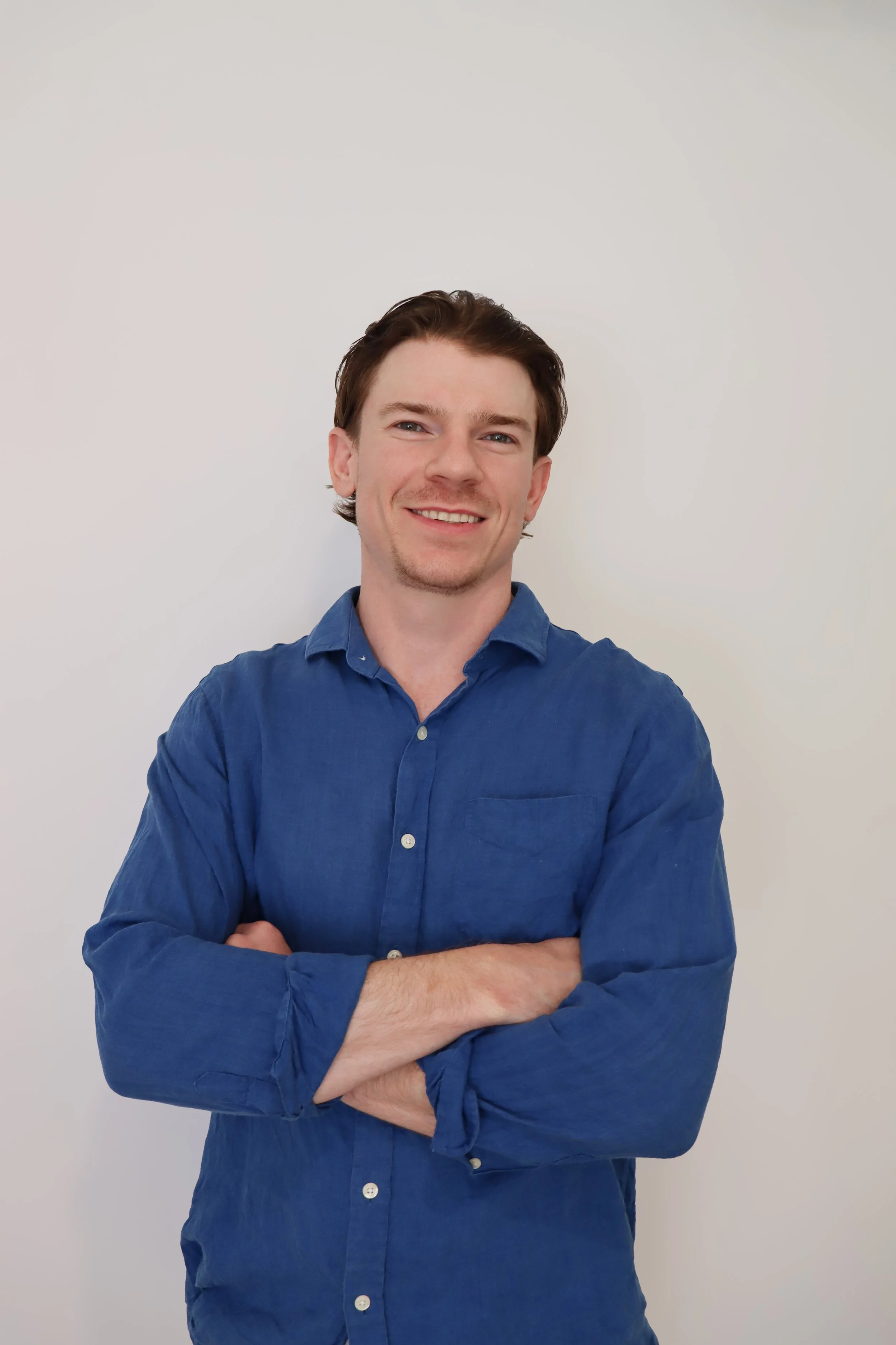 A young man with brown hair, wearing a blue button-up shirt, standing with arms crossed and smiling against a plain white background.