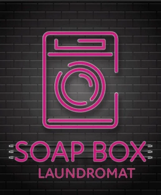 Neon sign with a pink washing machine outline and the words "Soap Box Laundromat" on a dark brick wall background.