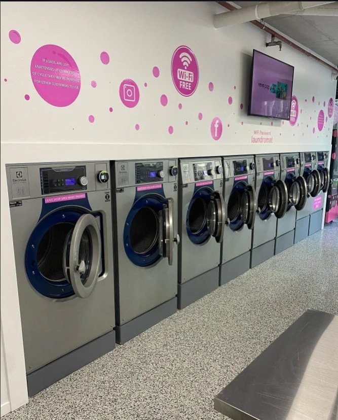 Row of front-loading washers in a laundromat with pink decor and a TV mounted on the wall.