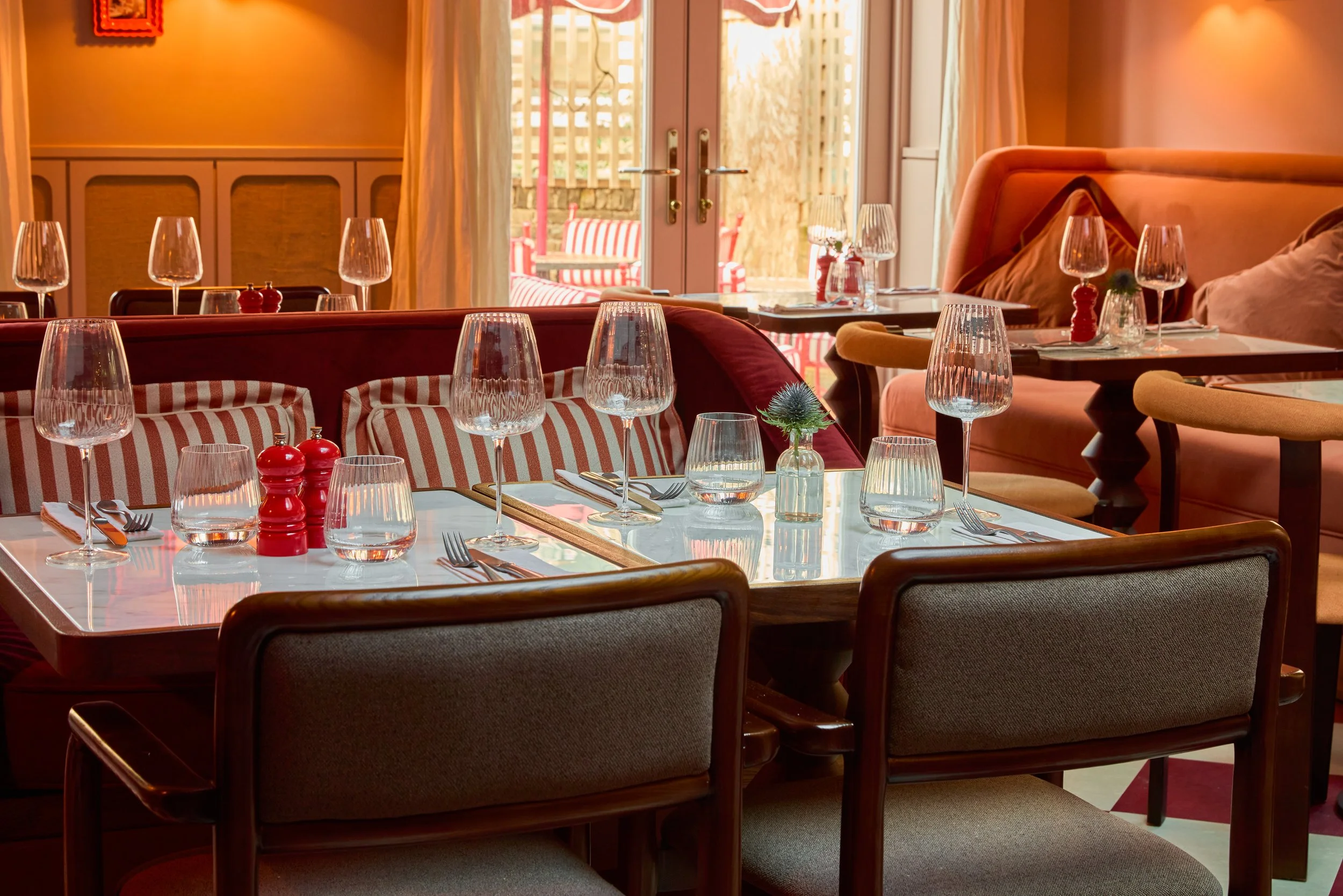 A cozy restaurant with a table set for dining, featuring wine glasses, water glasses, red pepper mills, a small plant, and utensils, with a view outside through glass doors.