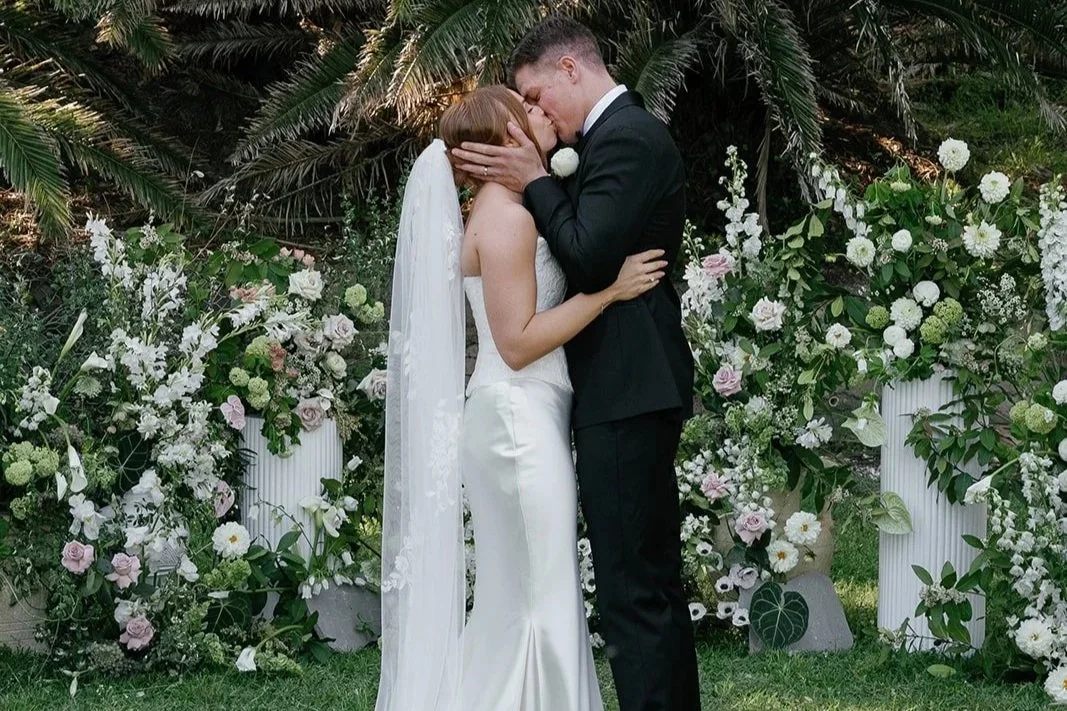 A bride and groom kiss in front of floral wedding decor featuring white and pink flowers, tall white vases, and lush greenery.