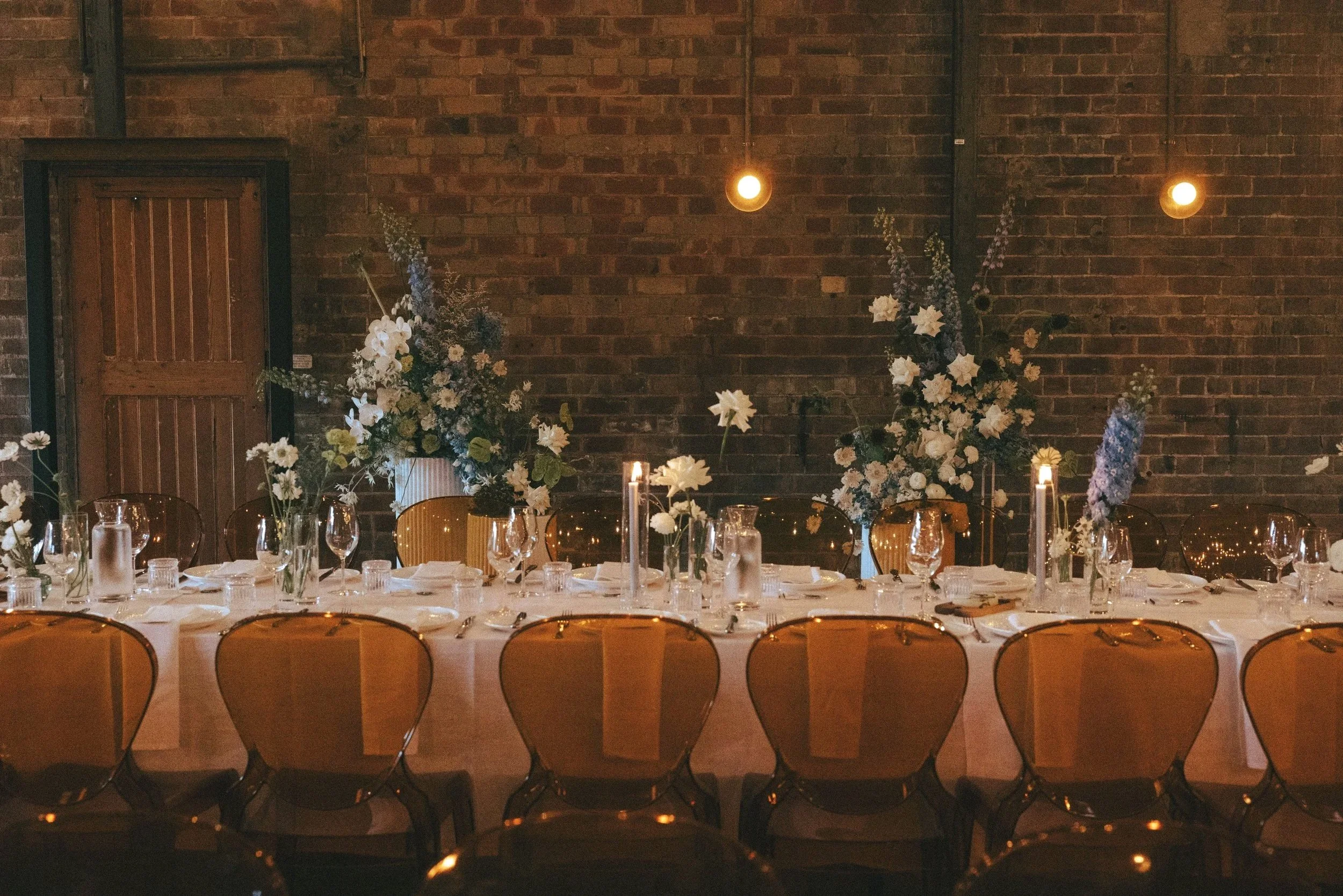 Elegant dining table setup for a special event with floral centerpieces, candles, and glassware against a brick wall with hanging lights.