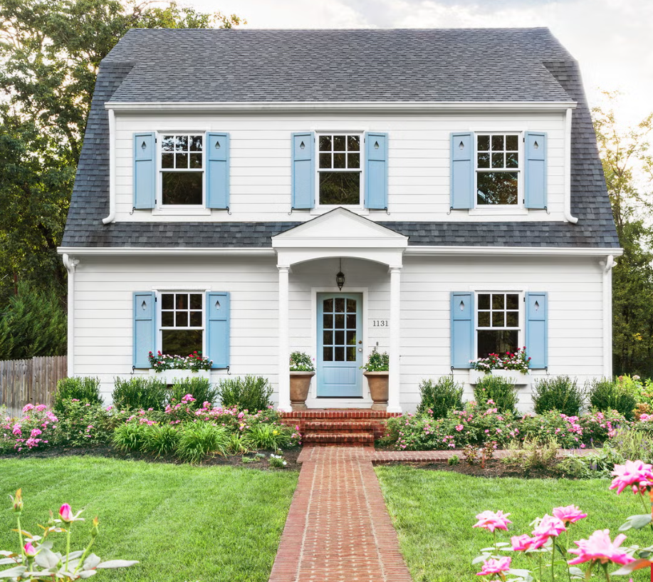 White two-story house with light blue shutters, front porch with white columns, and brick pathway leading to the front door, surrounded by a garden with pink flowers and green grass.