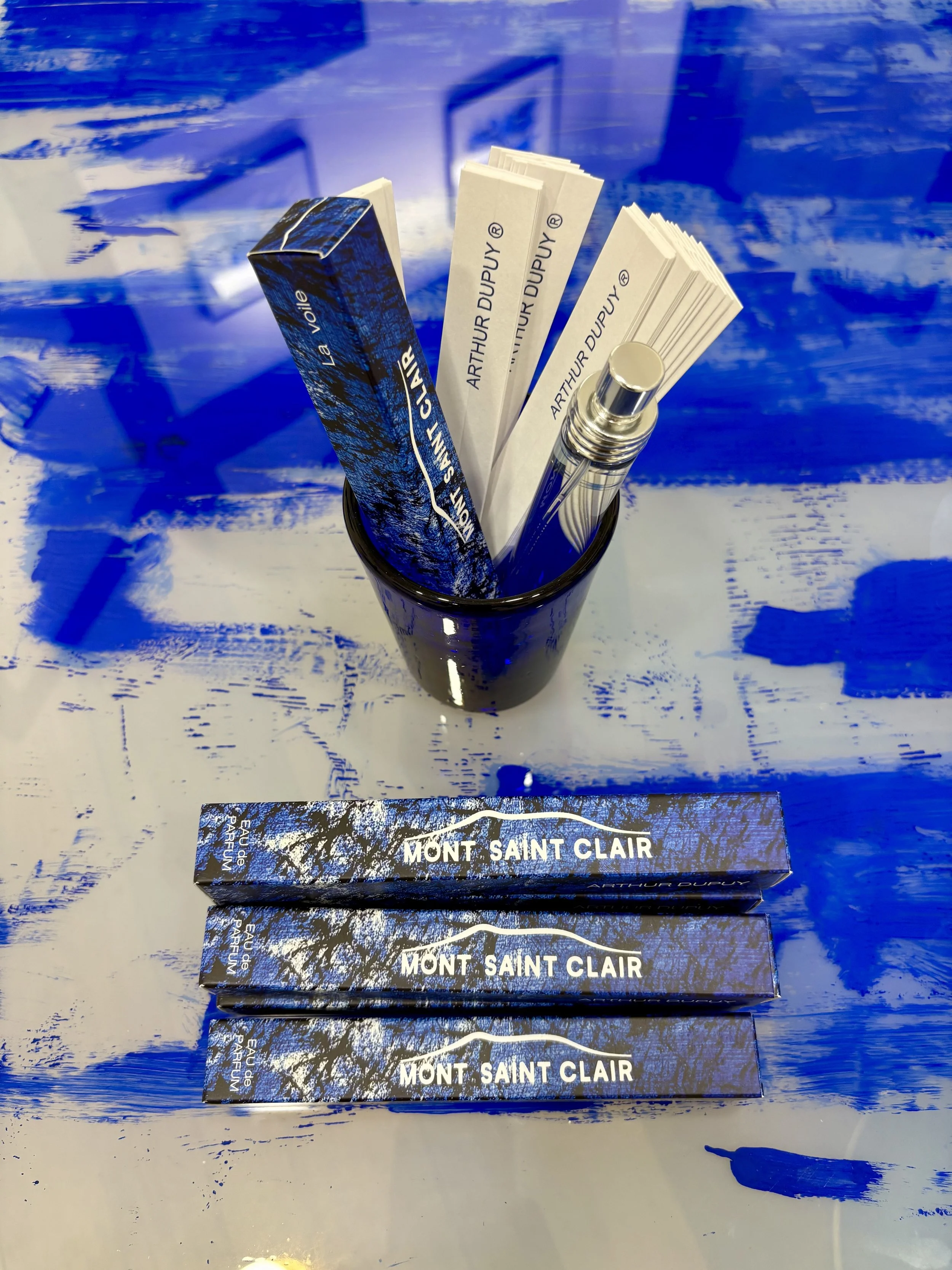 Boîte d'Eau de Parfum par Arthur Dupuy, Mont Saint Clair et un stylo dans un vase contenant plusieurs papiers avec le nom Arthur Dupuy, avec un fond bleu et blanc.