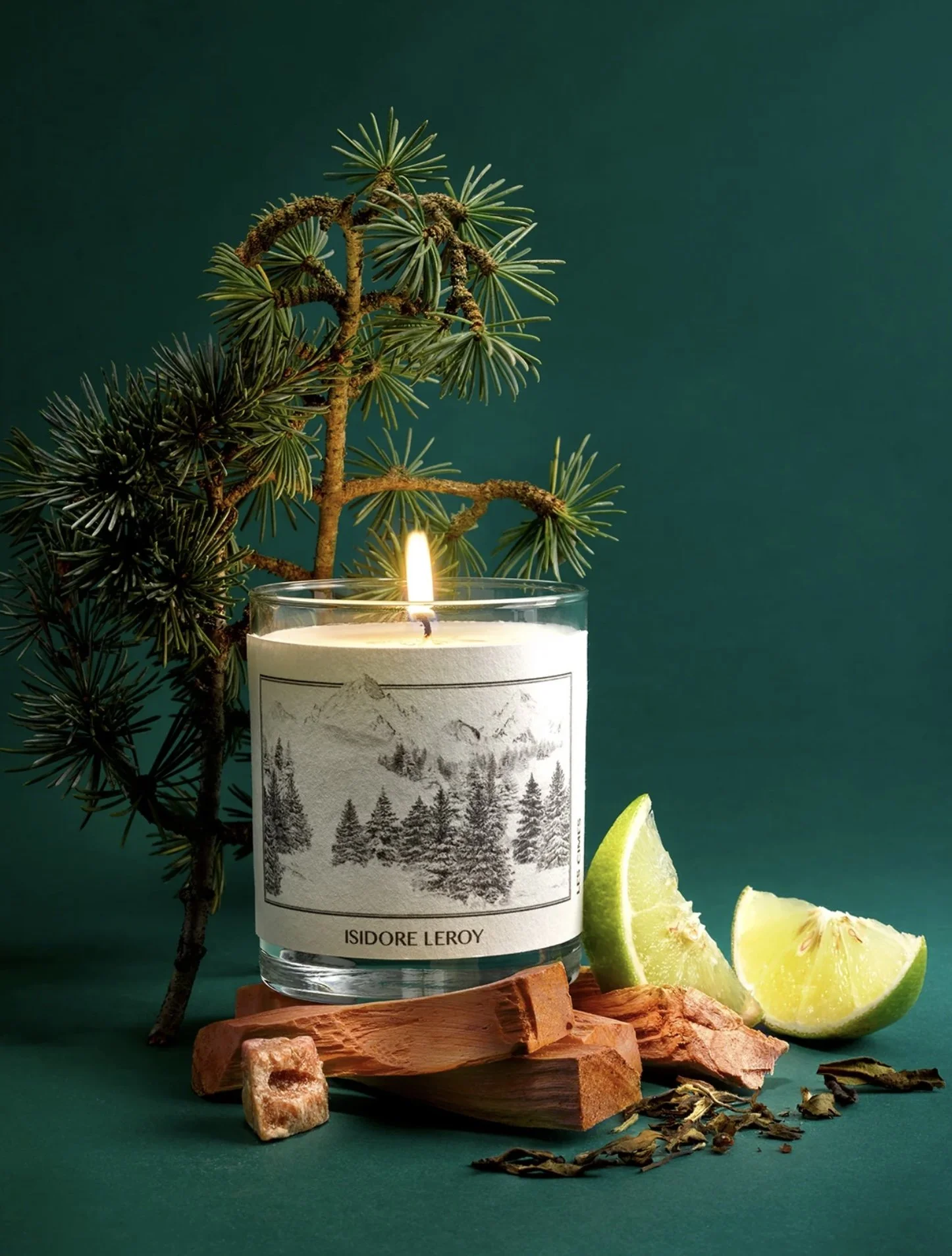 Bougie parfumée avec une étiquette de paysages enneigés, deux quartiers de citron vert, morceaux de bois, petits morceaux d'herbes séchées, et une branche de pin, sur un fond vert.
