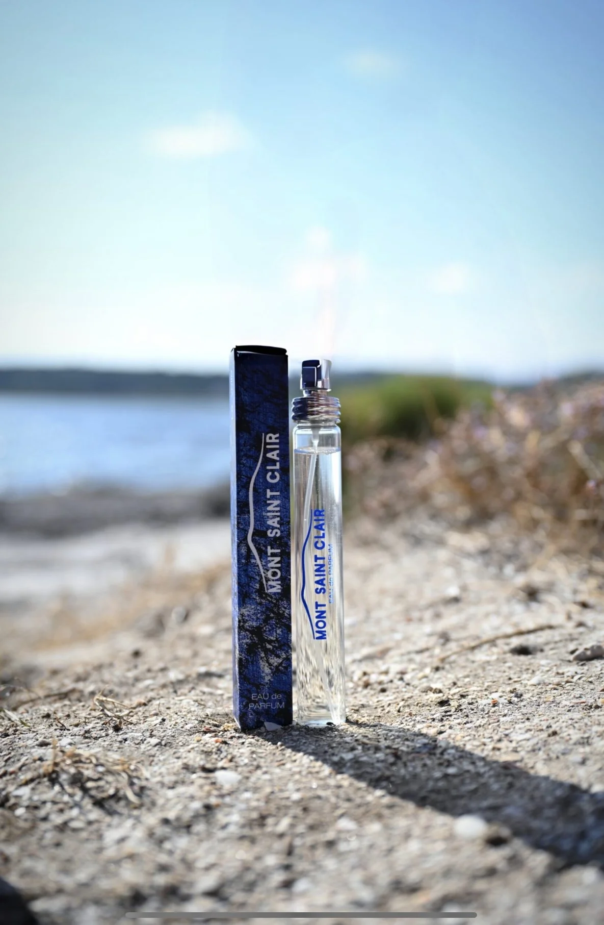 Un flacon de parfum créé par Arthur Dupuy, Mont Saint Clair et son emballage, posés sur le sol près d'une plage, avec la mer en arrière-plan et un ciel bleu.