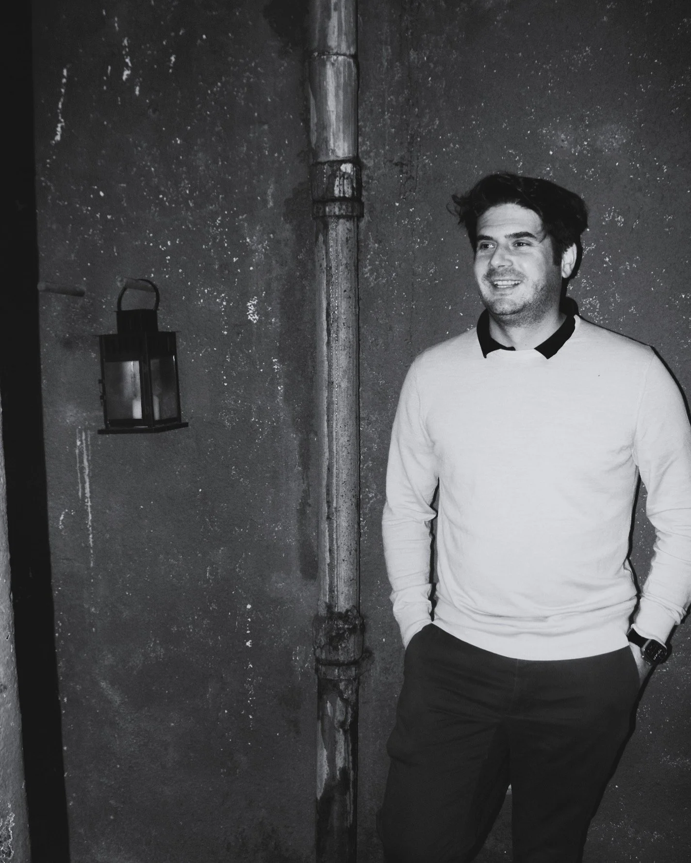 Portrait d'Arthur Dupuy, portant un pull blanc avec col noir, souriant, debout avec une main dans la poche, dans un endroit sombre avec un mur en béton et une lanterne suspendue.