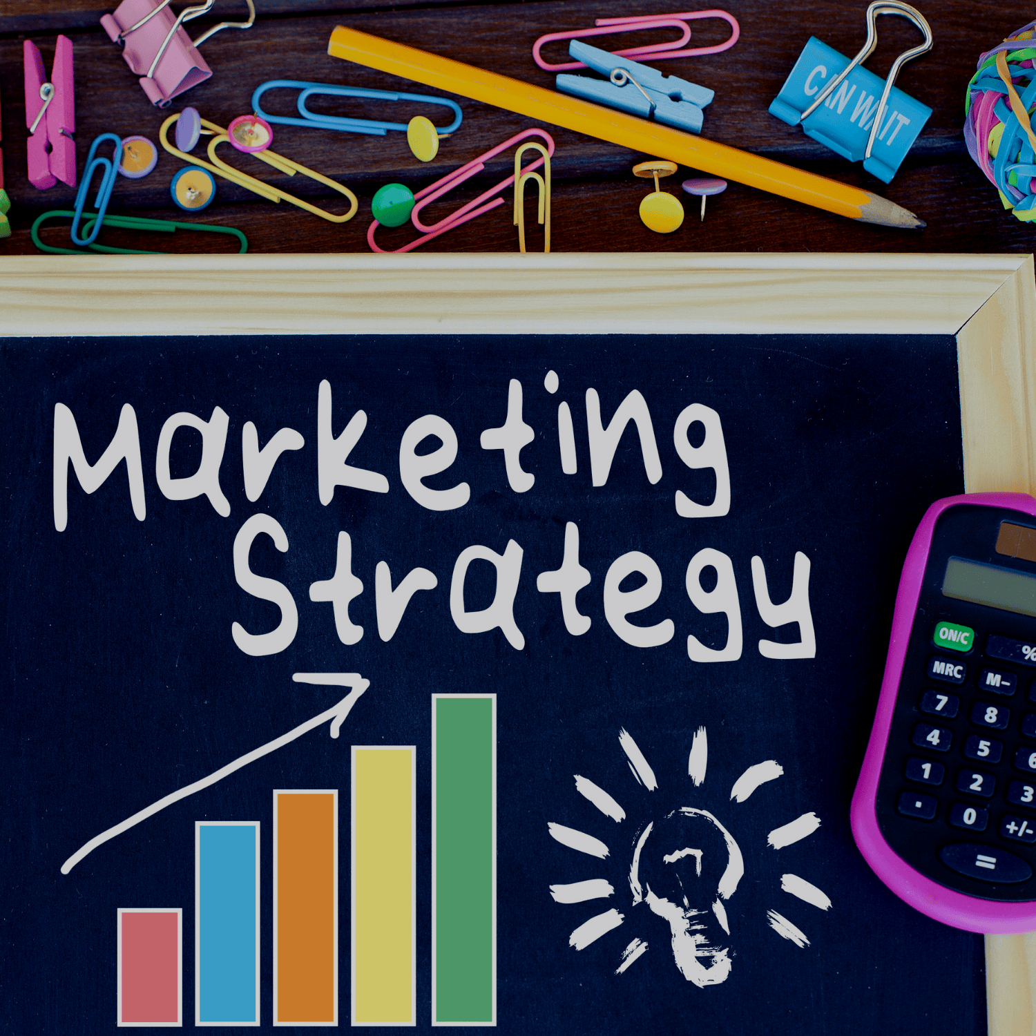 A blackboard with the words 'Marketing Strategy' written on it. A colorful bar chart with an upward arrow, a white lightbulb drawing, and various office supplies like paper clips, push pins, a pencil, and a calculator are on a wooden surface above the blackboard.