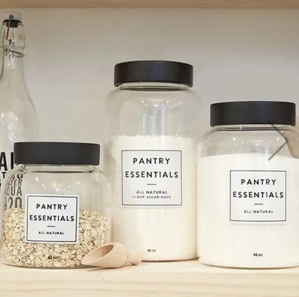 Three clear glass jars with black lids labeled 'Pantry Essentials All Natural', containing oats, flour, and sugar, on a white shelf with a wooden scoop and a bottle in the background.