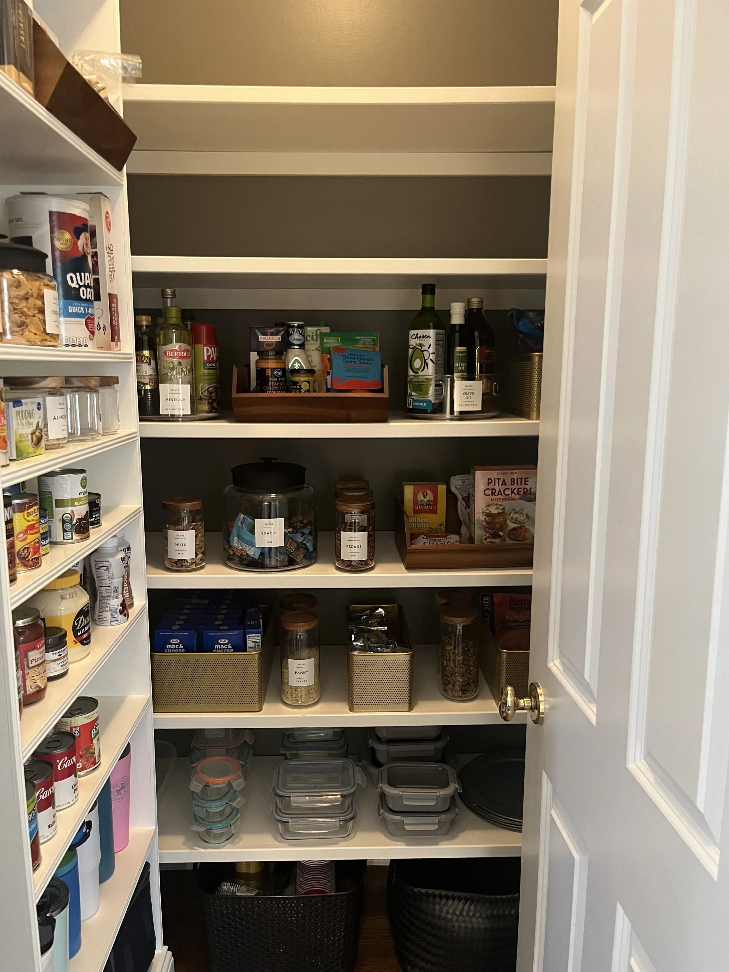 Pantry with shelves containing jars, boxes, bottles, and containers of food items, spices, and snacks, with a door partially open on the right side.