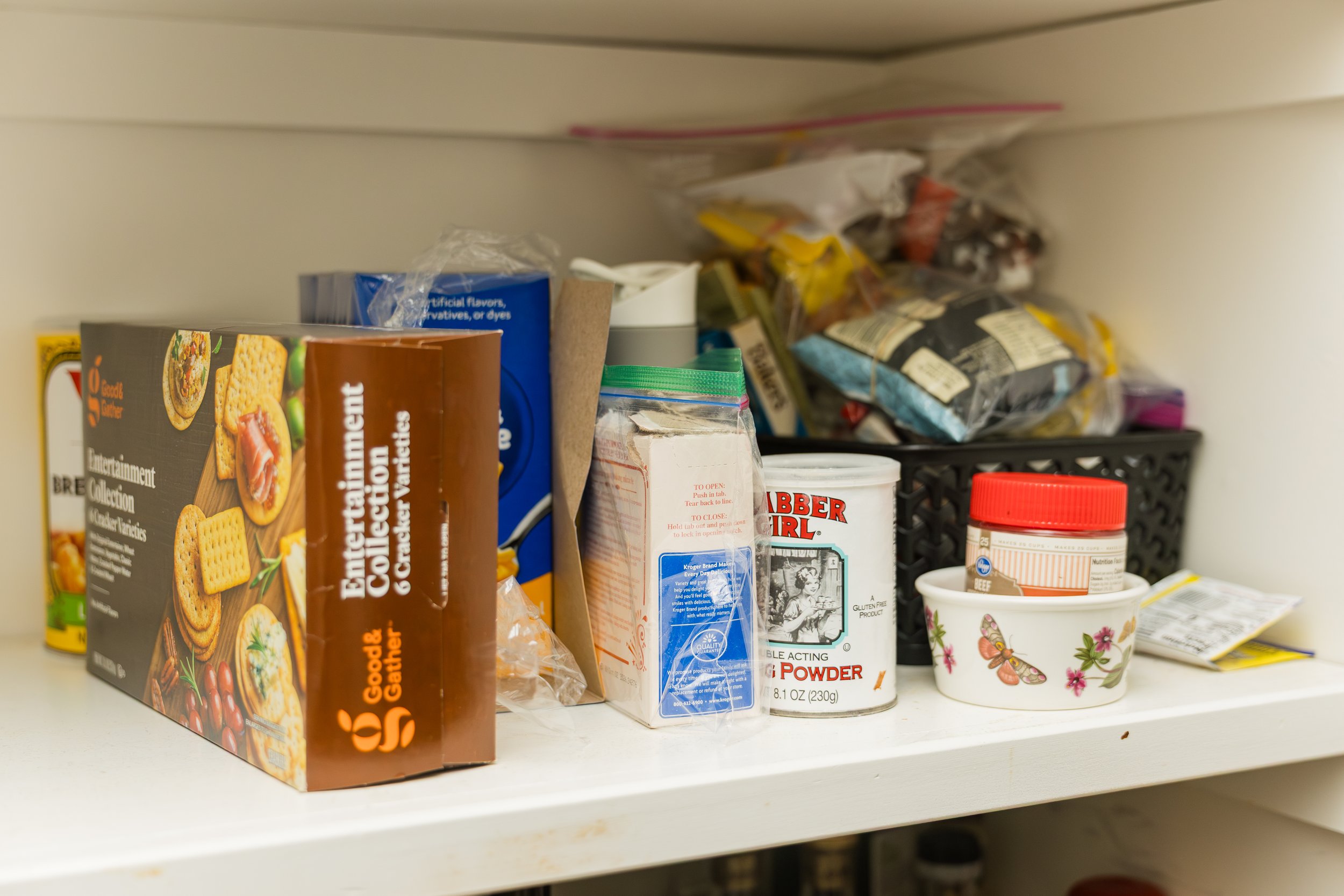A pantry shelf filled with various food items including boxed crackers, canned goods, a jar, a container of baking powder, and small bowls, with some items stored in plastic bags and a black plastic basket.
