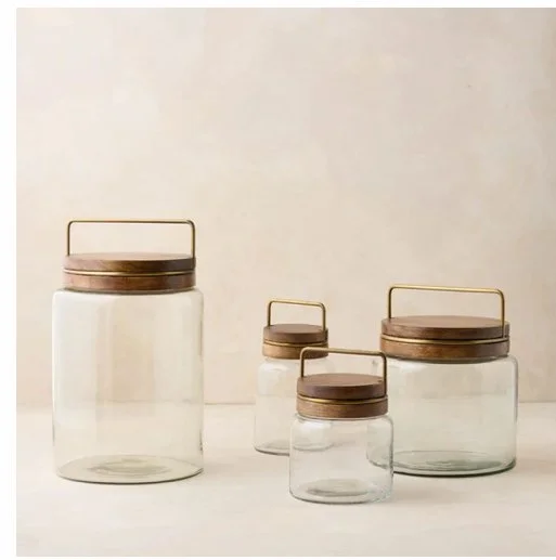 A set of four clear glass jars with wooden lids and metal handles, varying in size, arranged on a neutral surface.