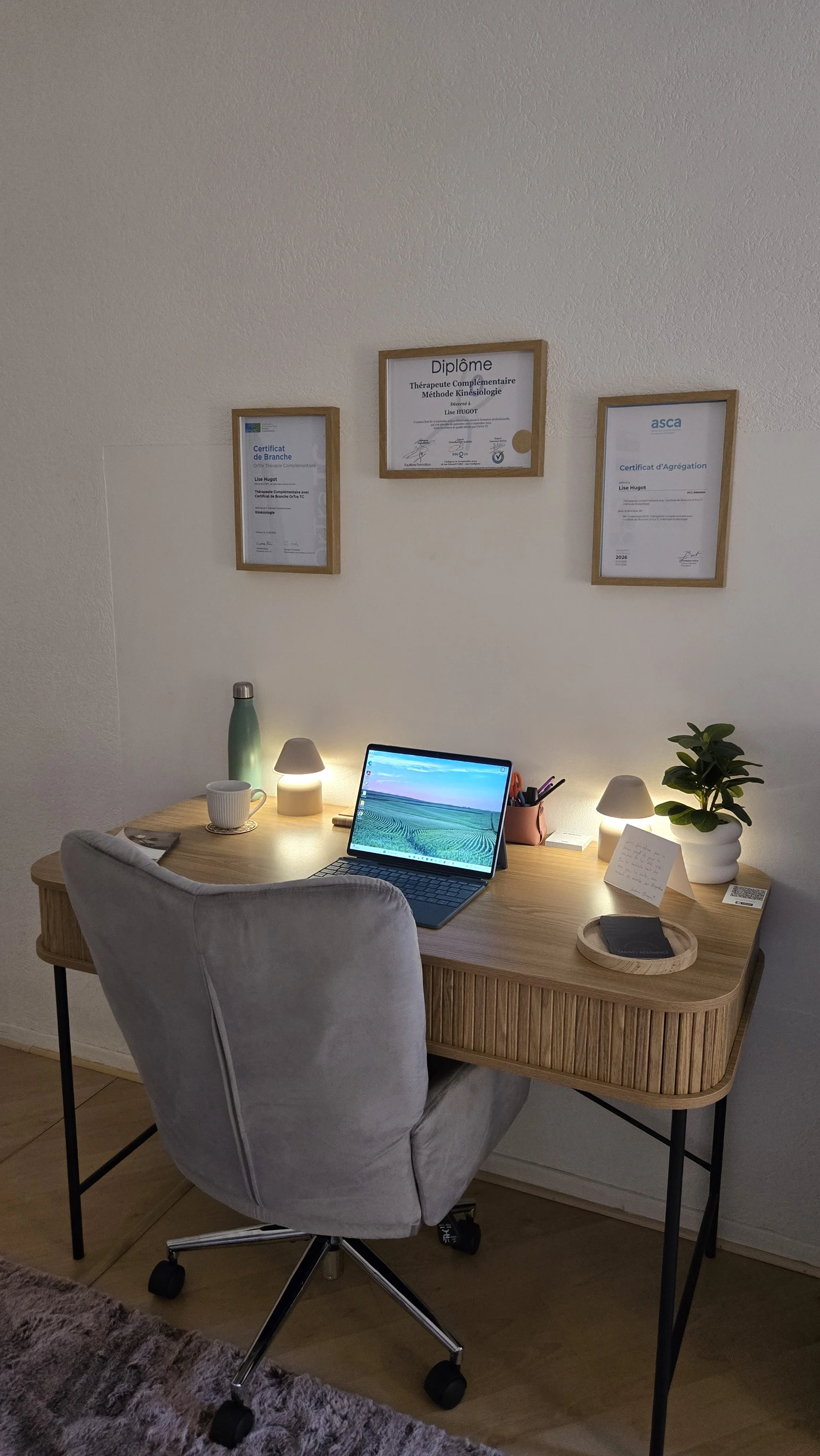 Un bureau avec un ordinateur portable, une tasse, des lampes, un vase avec une plante, des notes manuscrites, et des diplômes encadrés au mur.