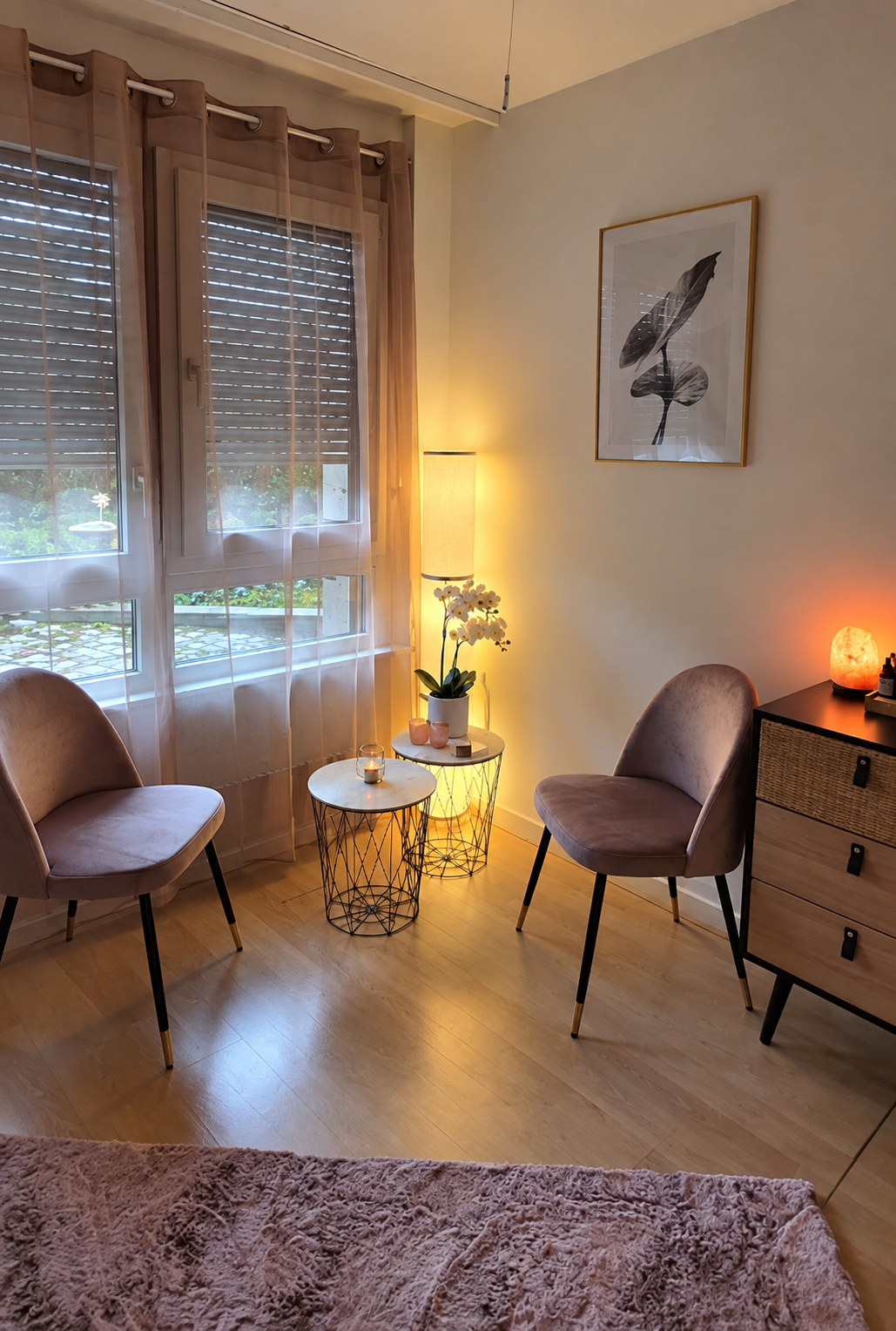 Une pièce de salon chaleureuse avec deux fauteuils rose clair, une petite table de bout en métal noir et marbre blanc, une plante orchidée et une lampe sur un tapis de pierre à côté d'une caisse de rangement. La pièce est éclairée par une lampe sur pied, une lampe de sel et la lumière naturelle venant des fenêtres avec rideaux translucides.
