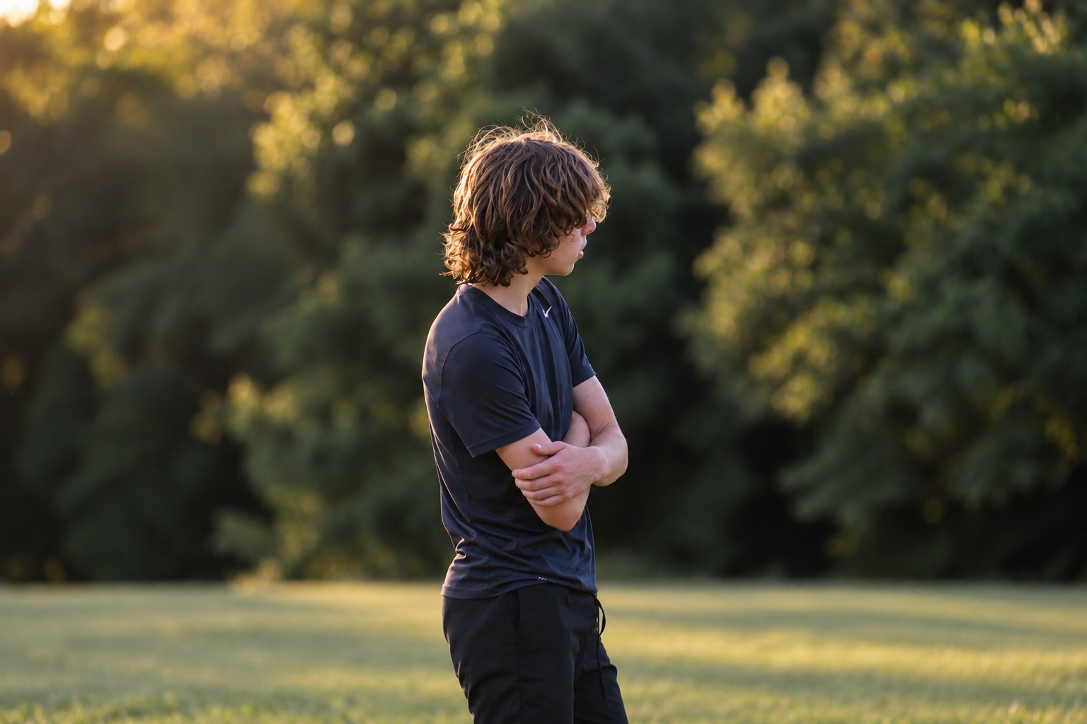 Jeune garçon aux cheveux curly, portant un t-shirt noir, debout dans un parc en plein air, avec les bras croisés, regardant à gauche, en fin d'après-midi avec un fond d'arbres.