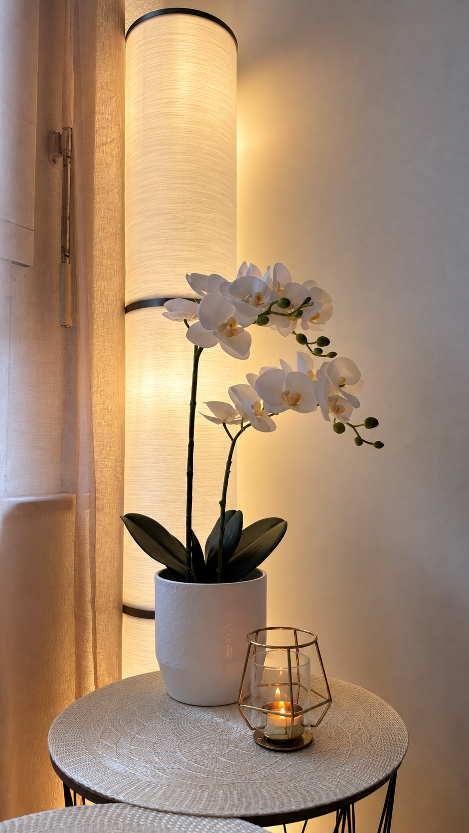 Une plante de orchidée blanche dans un pot blanc, posée sur une table ronde en rotin avec une bougie dans un récipient en verre avec une structure en métal doré, éclairée par une lampe cylindrique à lumière chaude.