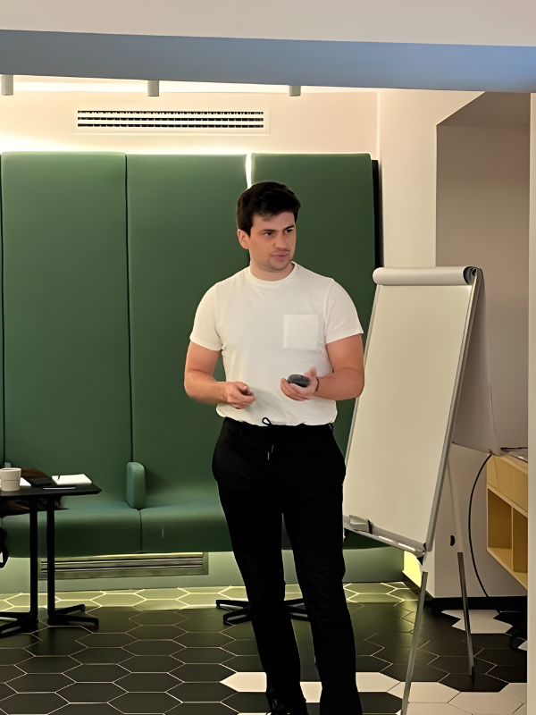 A young man in a white t-shirt and black pants standing in front of a whiteboard with a marker in hand, in a modern room with green seating and hexagonal floor tiles.