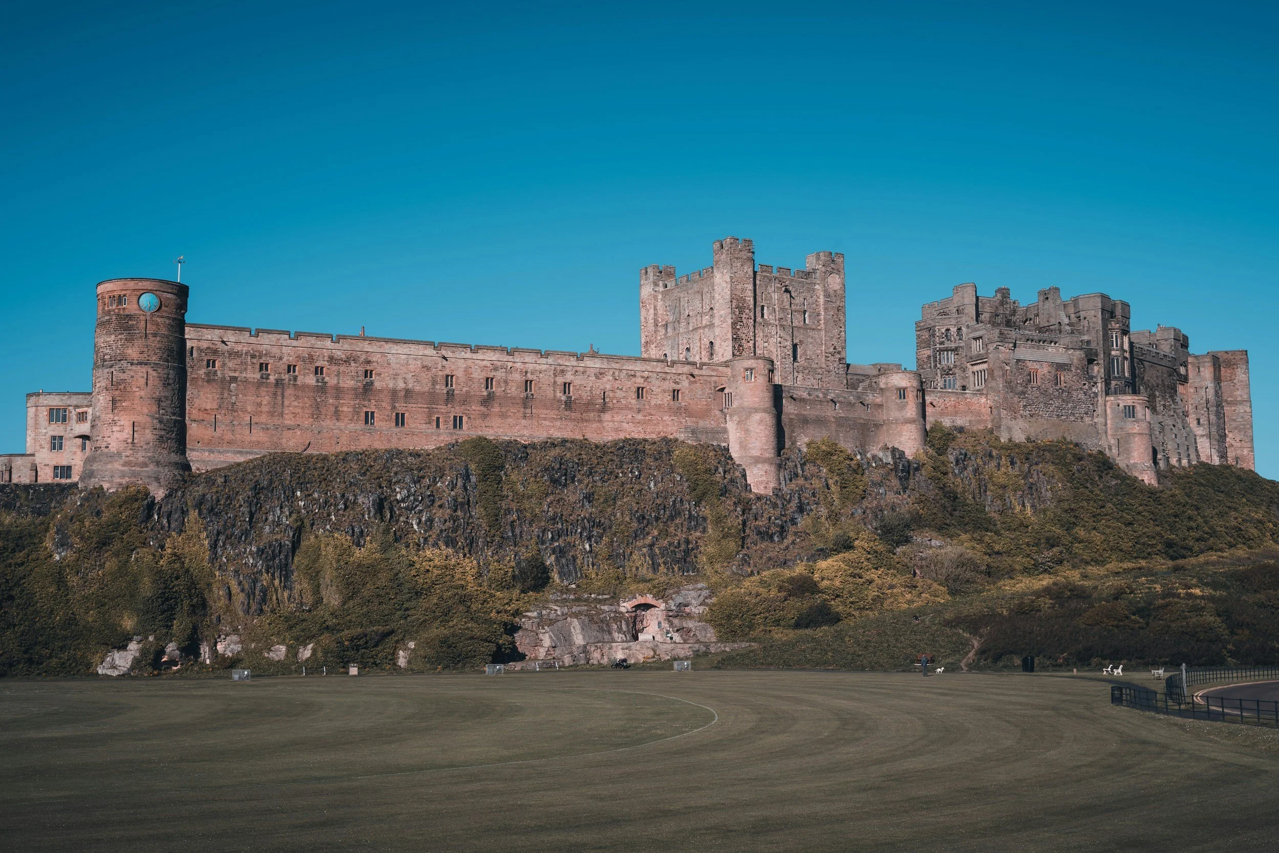 A large medieval castle with stone walls and towers sitting on a rocky hill, overlooking a grassy field under a clear blue sky.