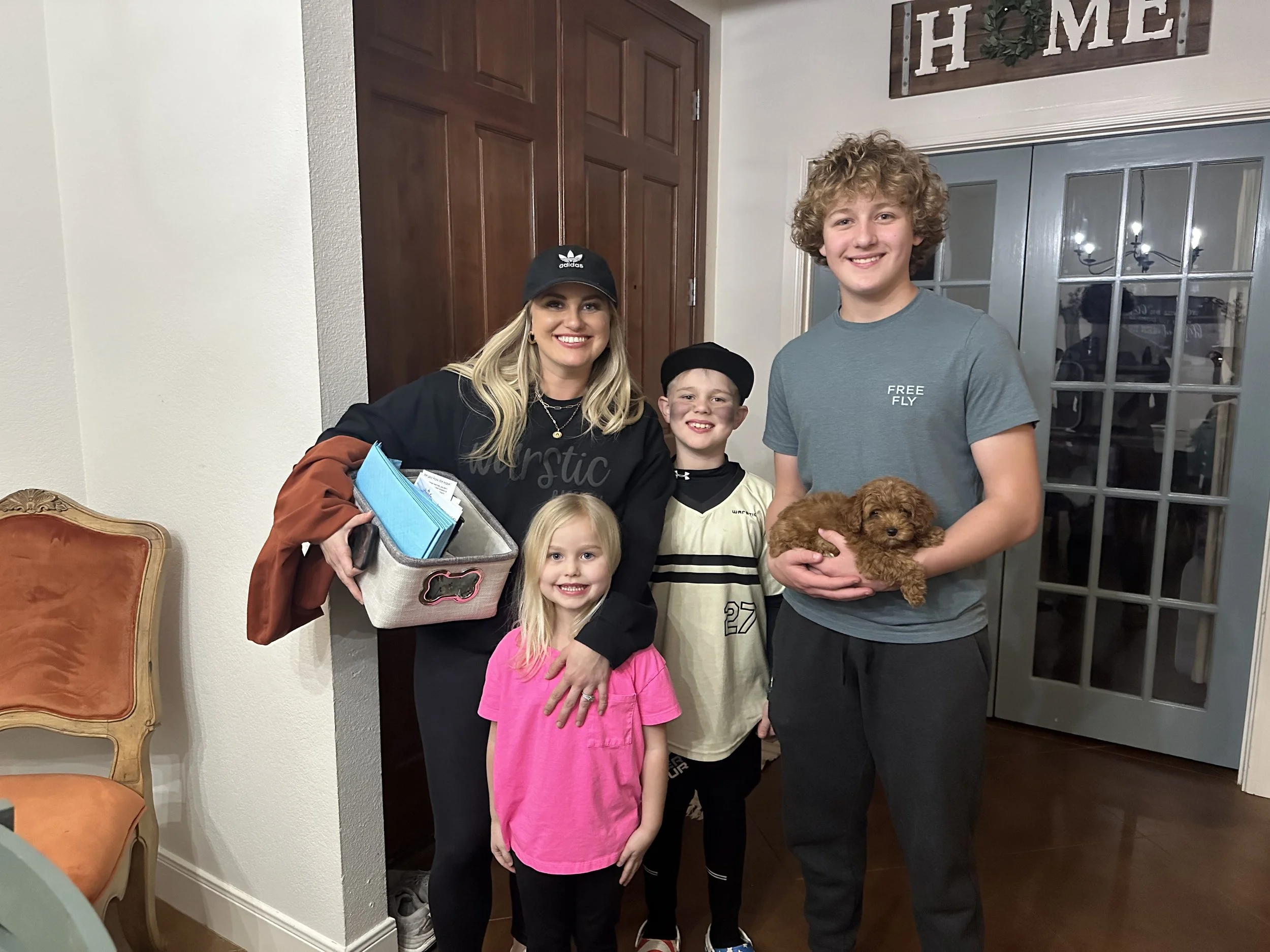 A group of five people, including children and adults, standing together indoors. One young man is holding a small brown puppy. They are smiling and posing for the photo, with a decorative wall sign that says "HOME" in the background.