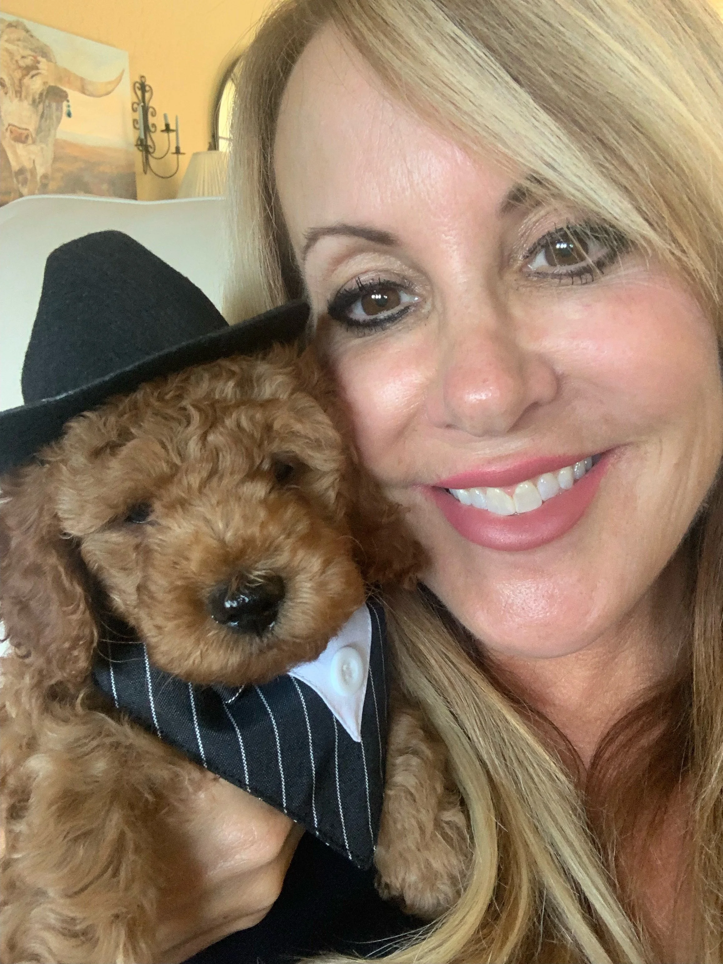 A woman with long blonde hair smiling and holding a small, curly-haired dog wearing a black hat and striped jacket.