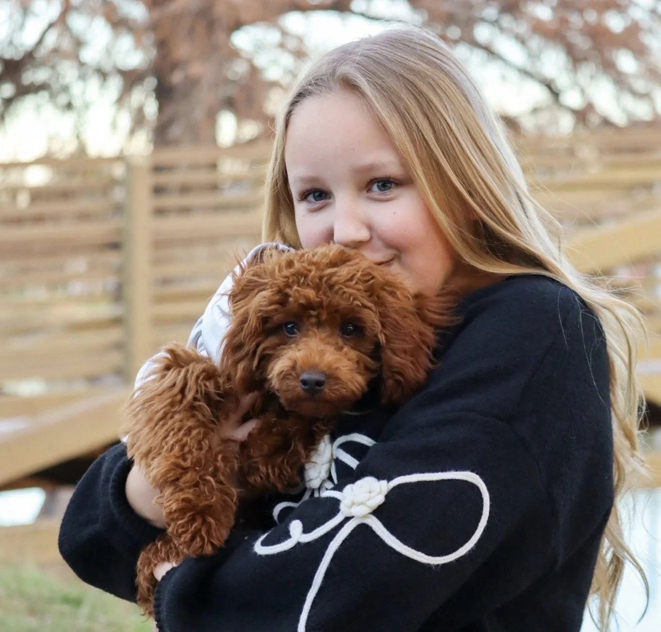 A young girl with long blonde hair holding a brown curly-haired puppy in her arms outdoors near a wooden fence, smiling at the camera.