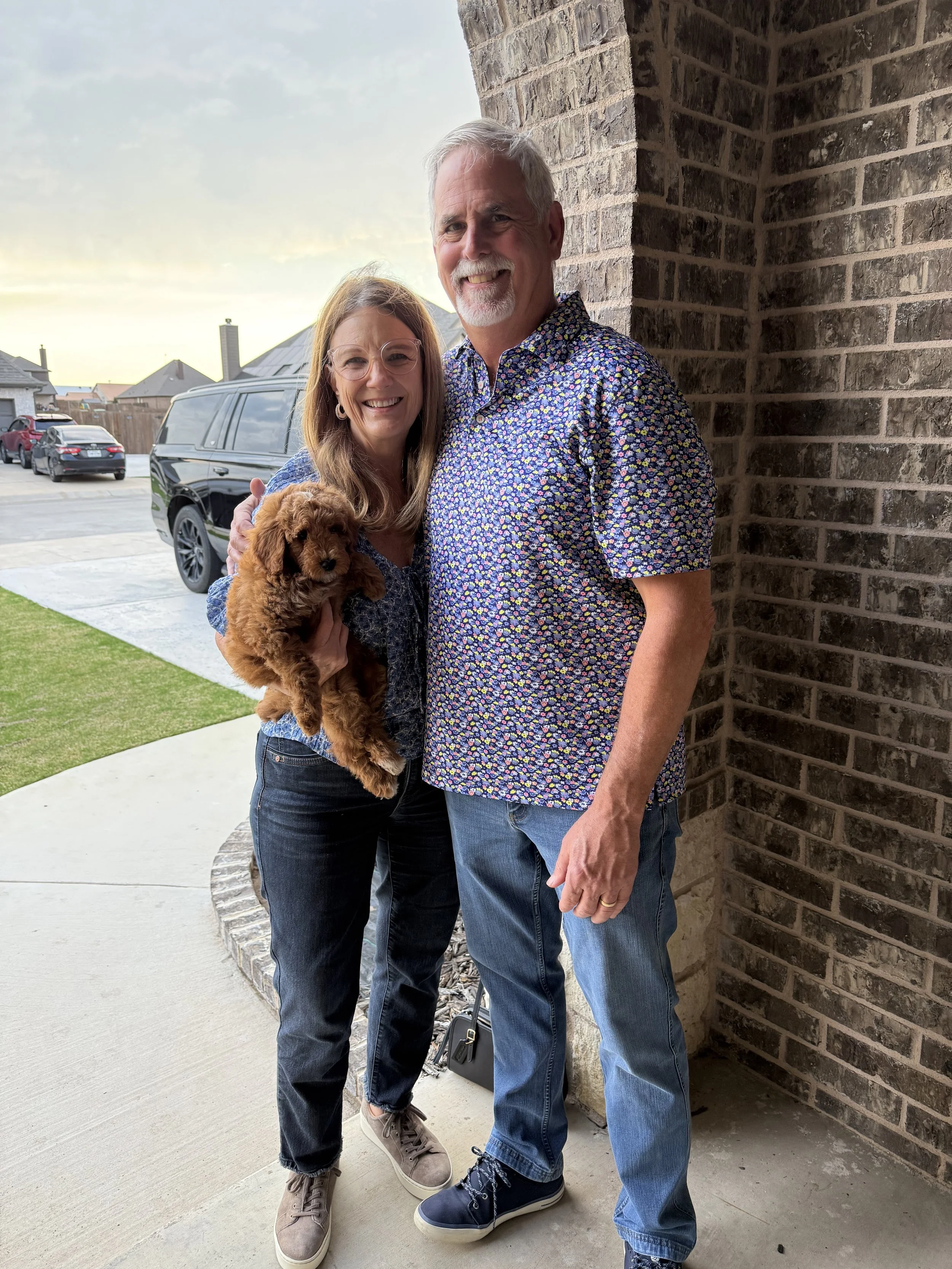 A smiling middle-aged man with gray hair and a beard, wearing a floral shirt, standing next to a smiling woman with glasses, long brown hair, wearing a patterned blouse, holding a small brown curly-haired puppy, standing outside near a brick wall, du