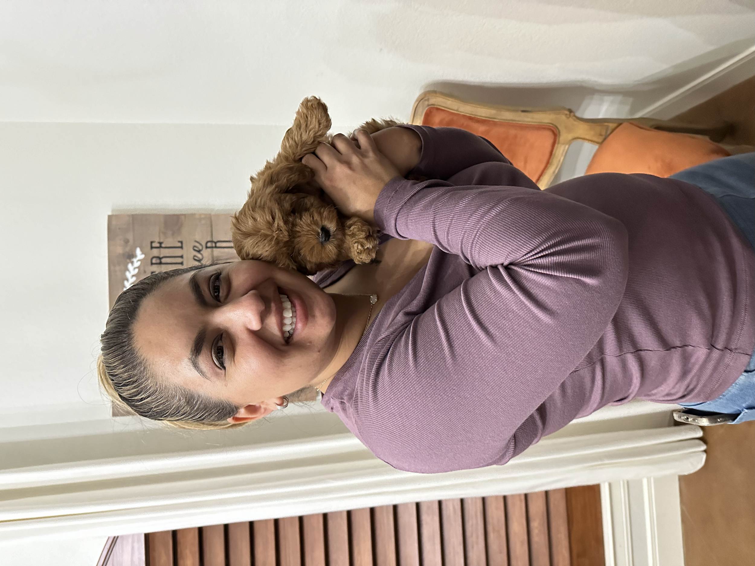 A woman holding a small brown puppy with curly fur, smiling at the camera, indoors with wooden furniture and a decorative sign on the wall.