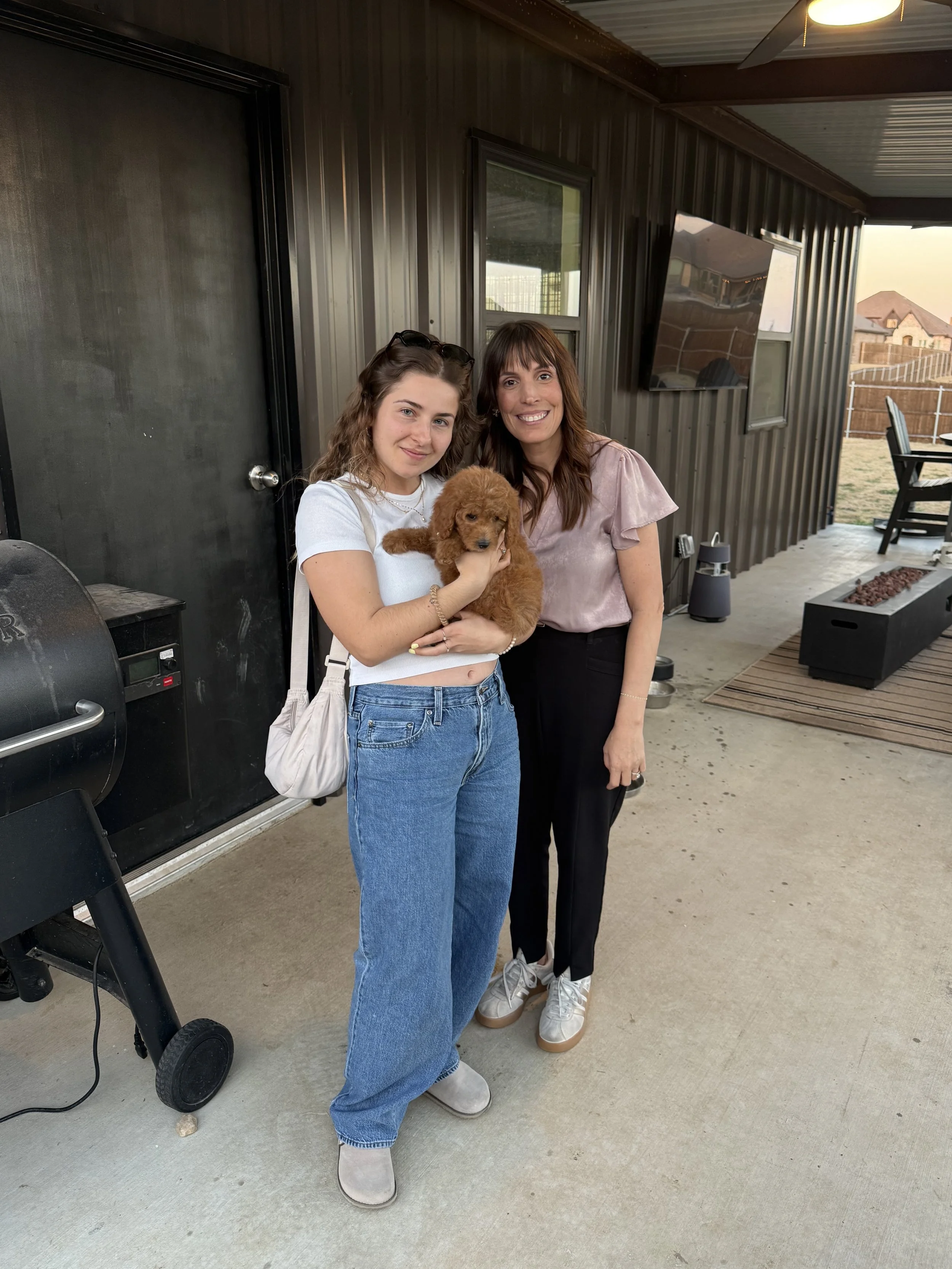 Two women standing on a porch, holding a small brown puppy. One woman is wearing a white shirt and jeans, while the other is in a pinkish blouse and black pants. The porch has a metal wall, a TV, a grill, and an outdoor fire pit.