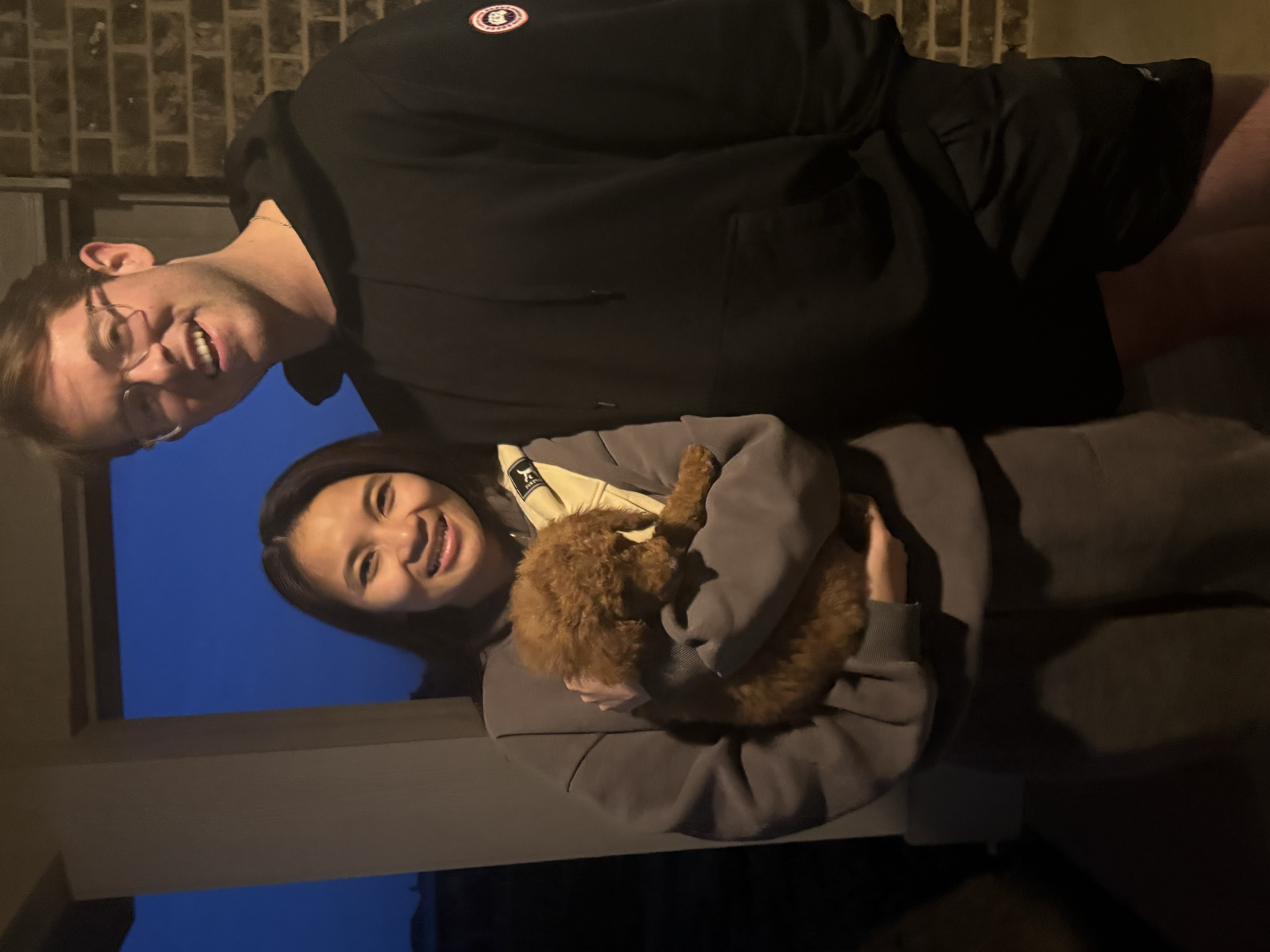 People are outside during dusk or evening with a brick wall behind them. One person is smiling and wearing glasses, another person is holding a small dog. The individuals are dressed in darker clothing, and the person with glasses has a patches or ba