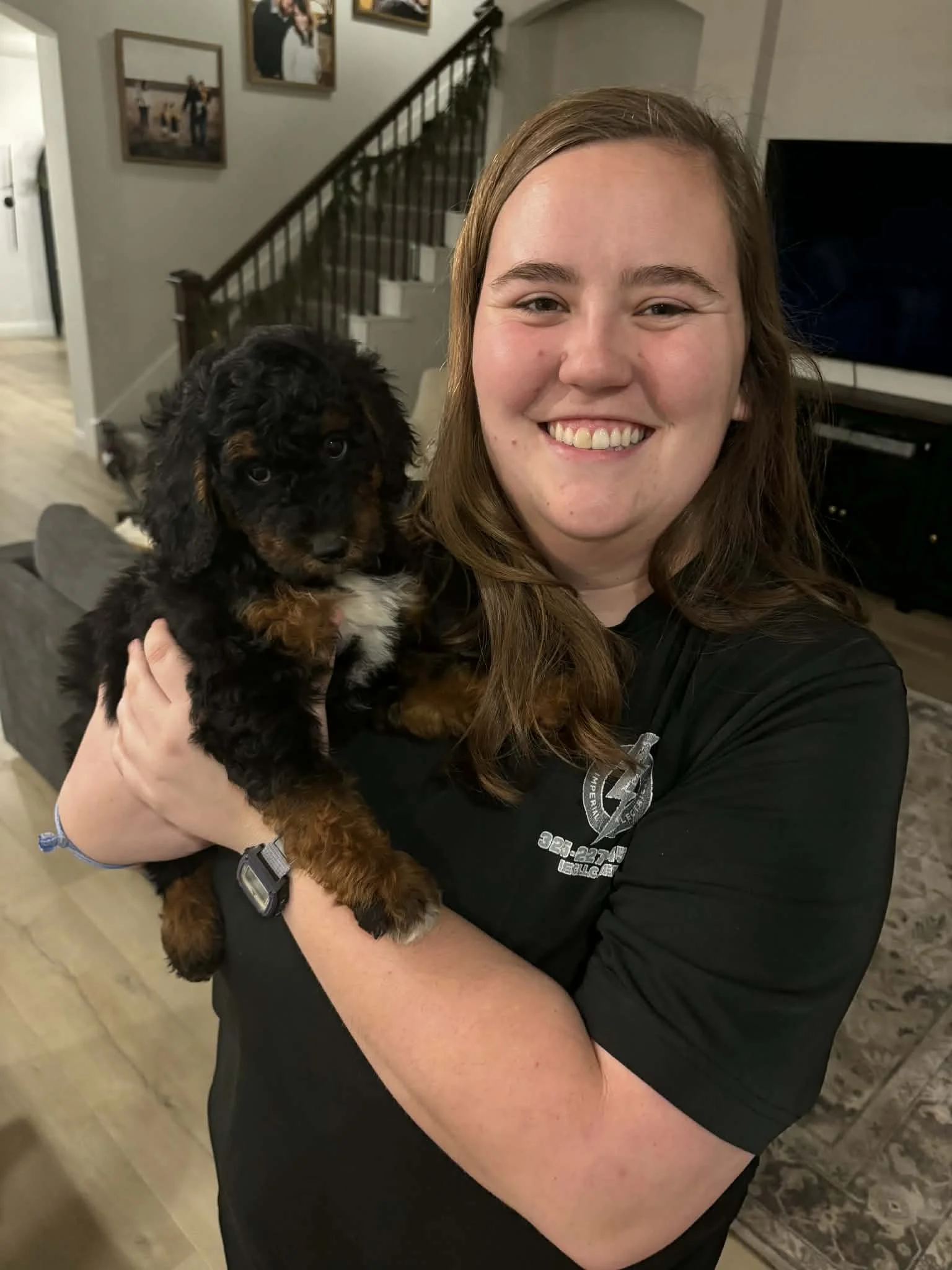 A smiling woman with long light brown hair holding a black and brown puppy, standing inside a home with framed pictures on the wall and a staircase in the background.