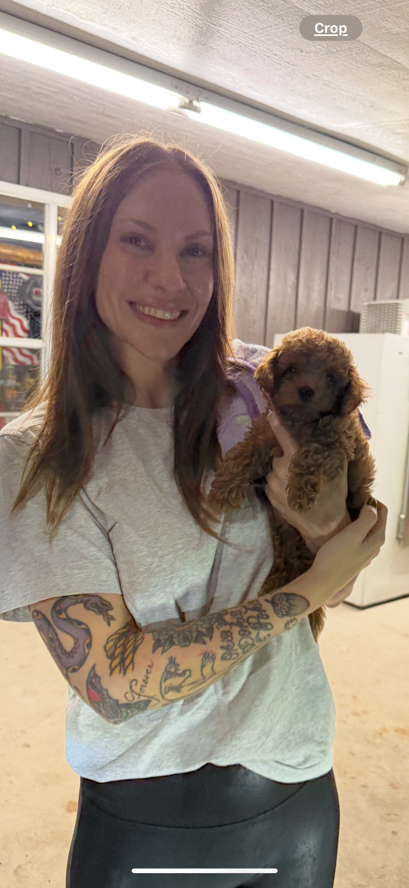 A woman with tattoos on her arm smiling while holding a small brown puppy in a room with wood-paneled walls and fluorescent lighting.