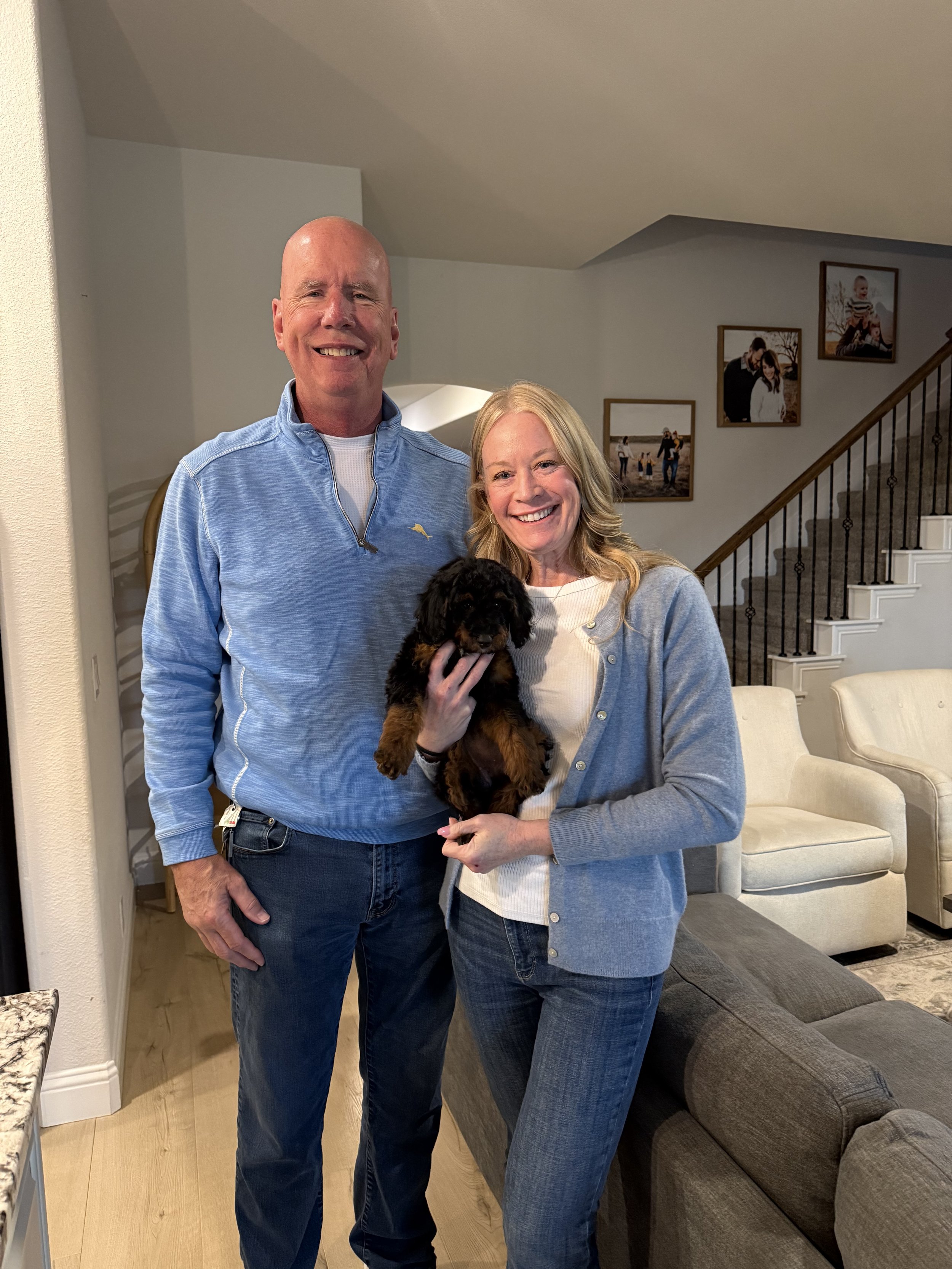 A smiling man and woman holding a black puppy in a living room, with photos hanging on the wall and a staircase in the background.