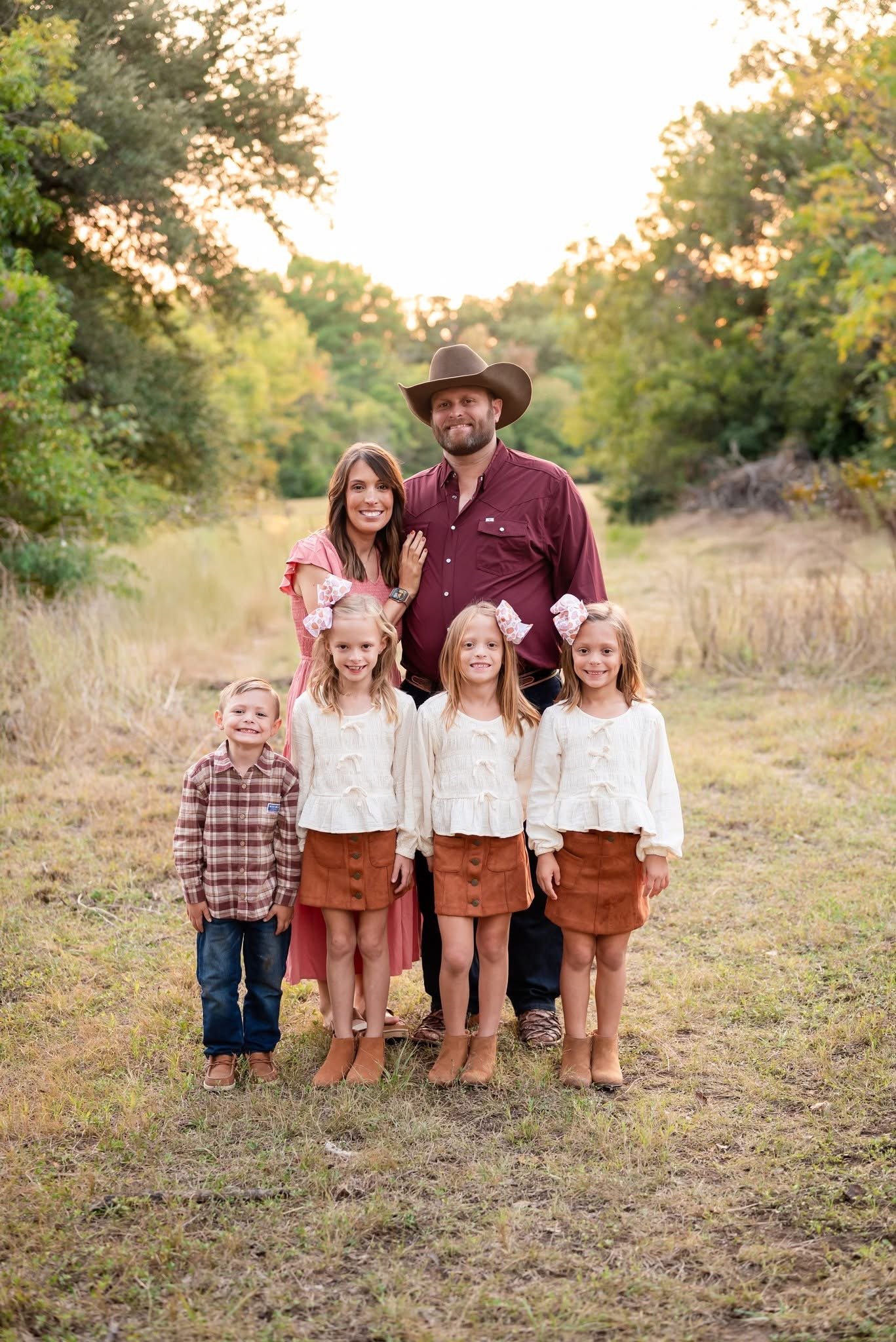 A family of six posing outdoors in a grassy clearing surrounded by trees with green and yellow leaves during the daytime. The father wears a cowboy hat and a maroon shirt, and the mother wears a pink dress. Four children stand in front: two girls with bow headbands and matching cream sweaters, and two boys with plaid shirts and brown skirts.