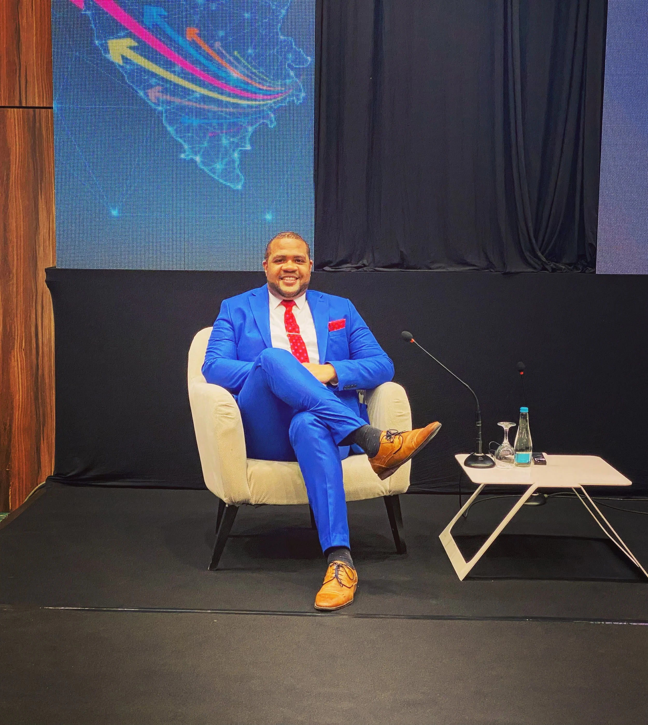 A man in a blue suit with a red tie sitting on a cream-colored armchair, smiling at the camera, with a wooden wall and a digital display showing colorful abstract graphics in the background.