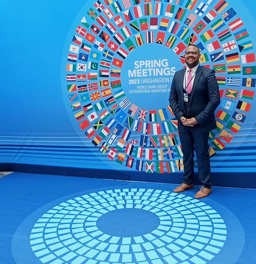 A man in a dark suit and brown shoes standing in front of a large circular display of various international flags at a 2023 Spring Meetings event in Washington D.C., with a blue background and floor design.